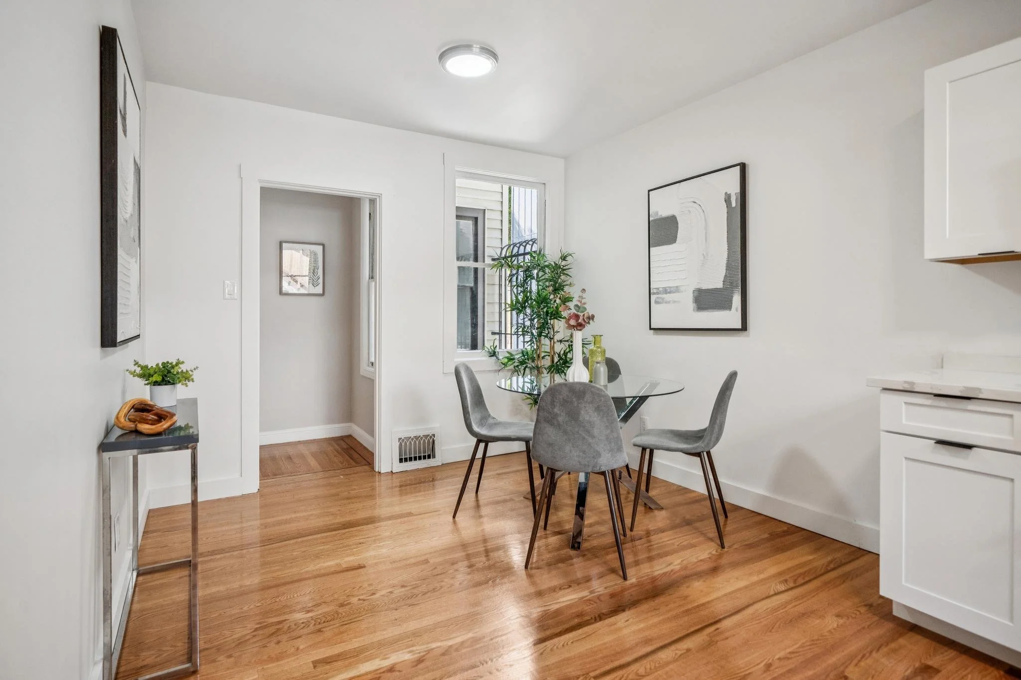A dining area with a glass table and four gray chairs, a window with a potted plant, framed artwork on the white walls, wooden flooring, and a glimpse of a kitchen to the right.