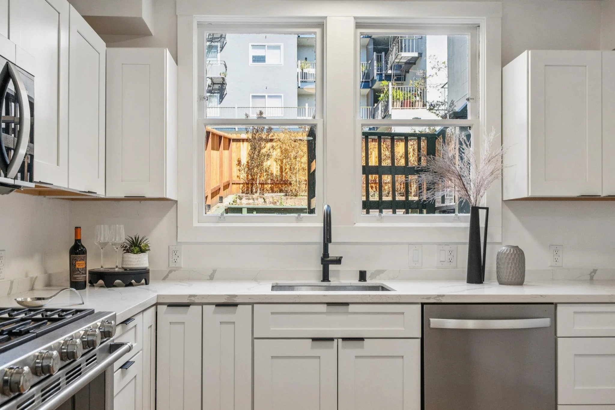 A modern kitchen with white cabinets, a marble countertop, and a large window above the sink showing an outdoor scene with buildings, balcony, and fence.