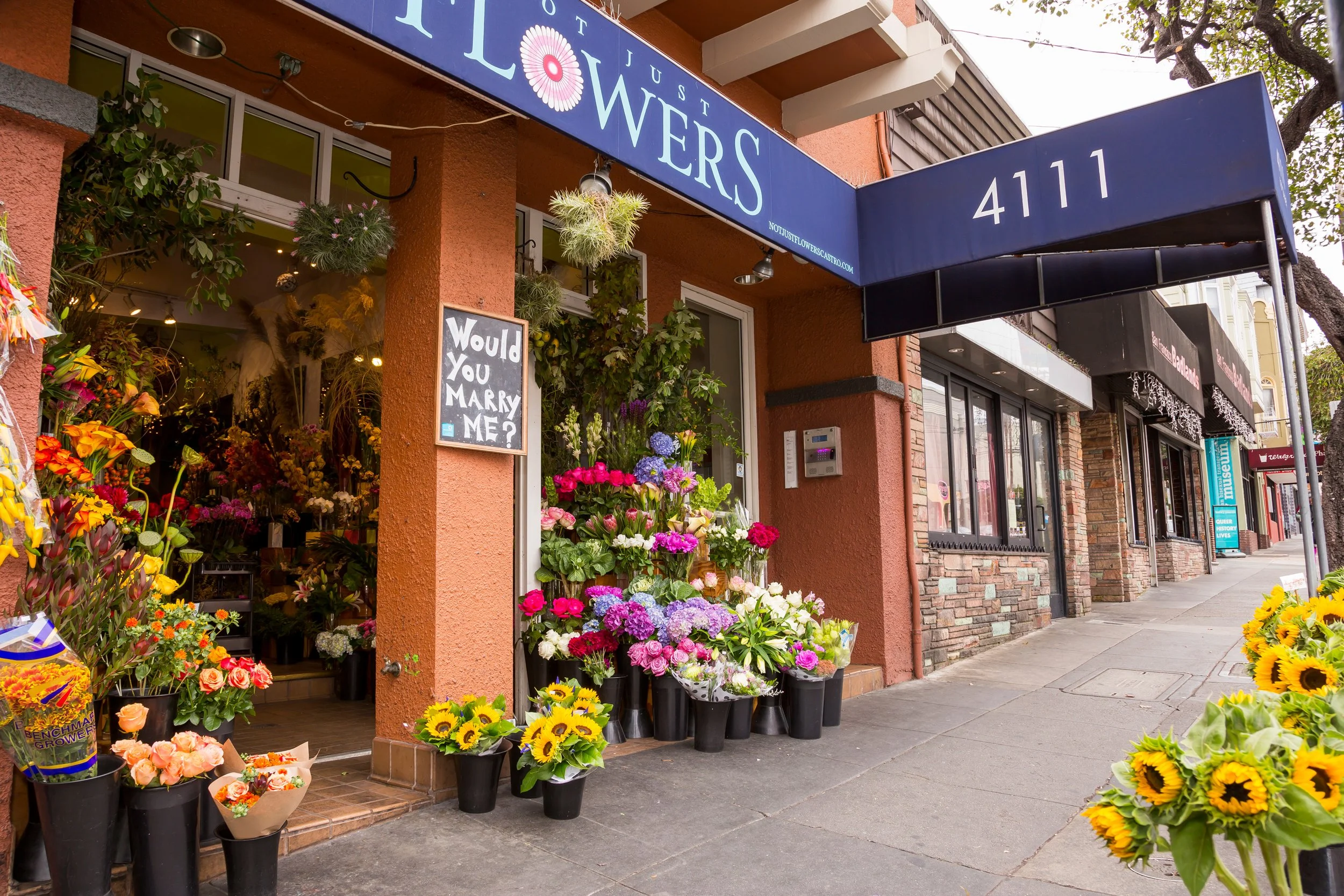 Flower shop storefront with a blue awning displaying number 4111, displaying colorful flowers including sunflowers, roses, and hydrangeas outside the shop, with a blackboard sign that says "Would You Marry Me?"