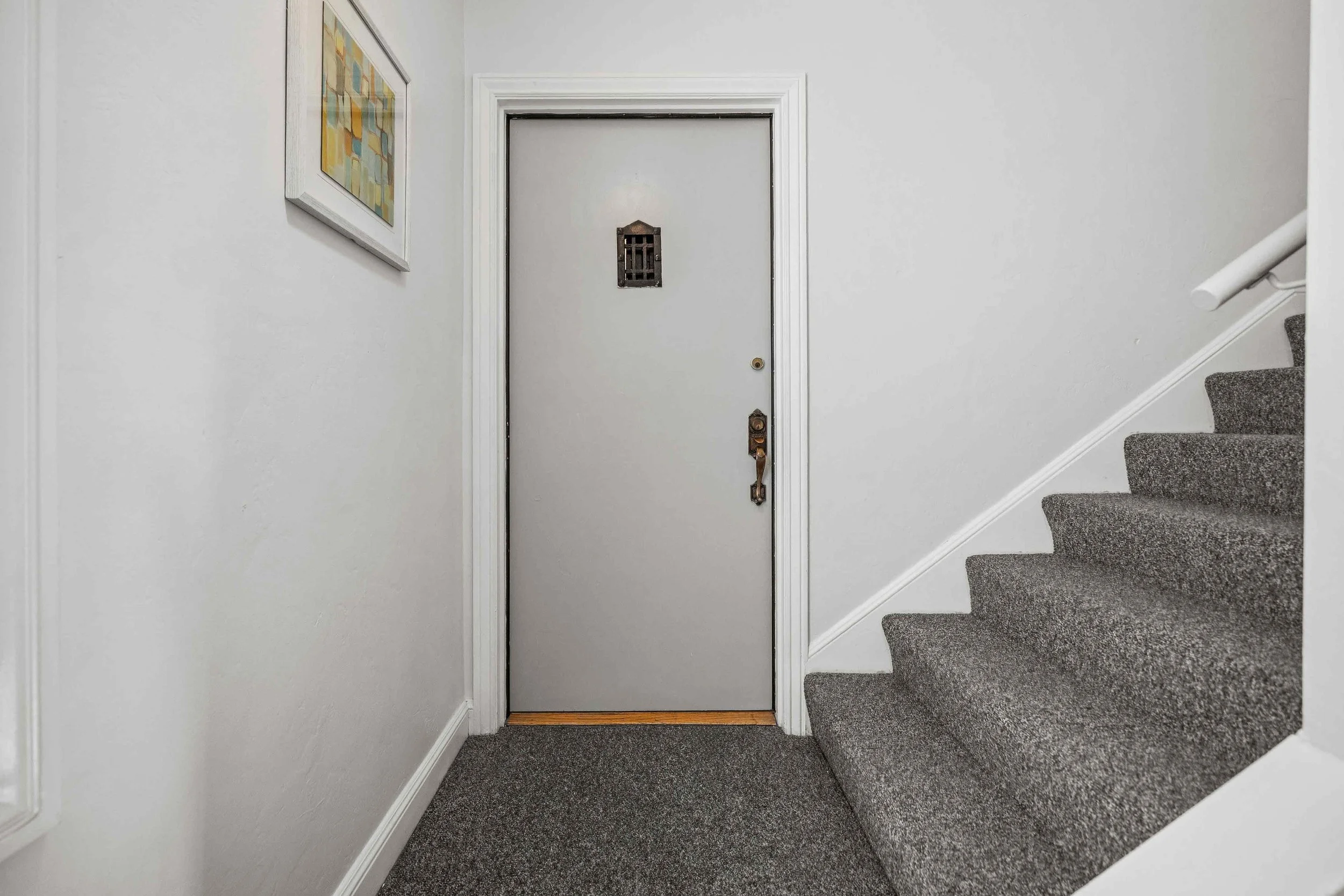 Interior of a residential staircase landing with a closed gray door and a staircase on the right side carpeted in gray, with a white handrail. There is a framed abstract art piece on the left white wall.