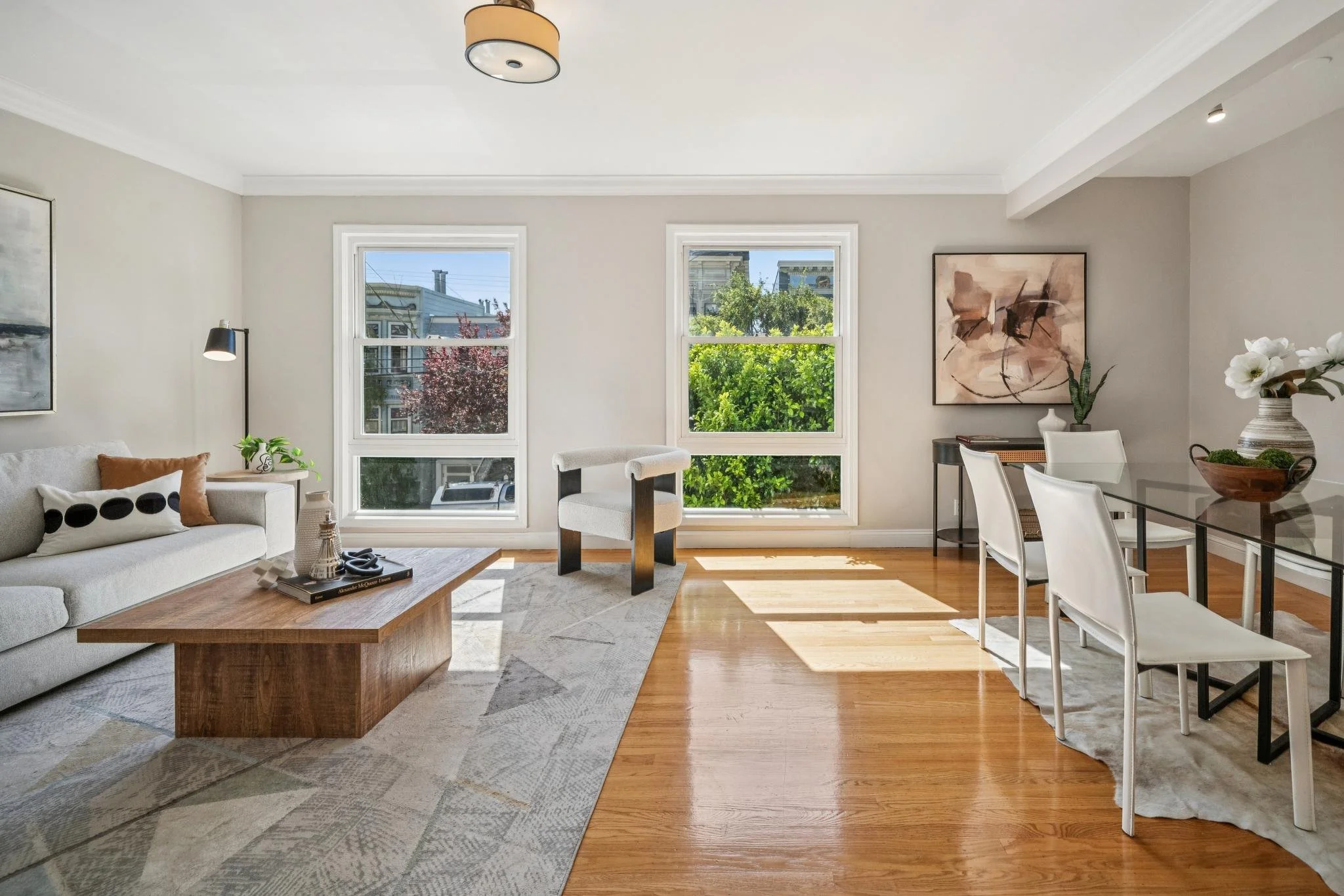 Bright living room with hardwood floors, beige sofa, wooden coffee table, abstract art on the walls, and large windows letting in natural light.