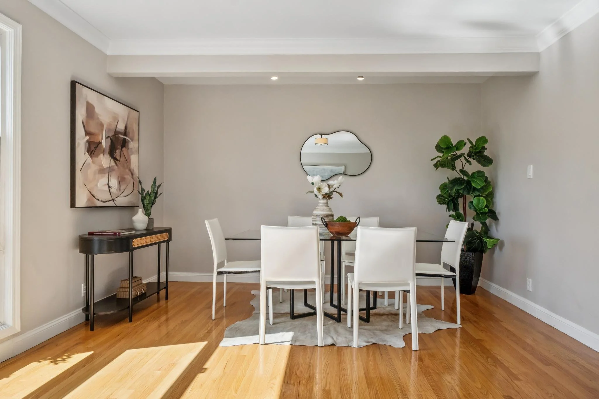 A modern dining room with a glass table, six white chairs, a wooden side table with a potted plant and books, a wall mirror, artwork, and a potted plant in a black vase.