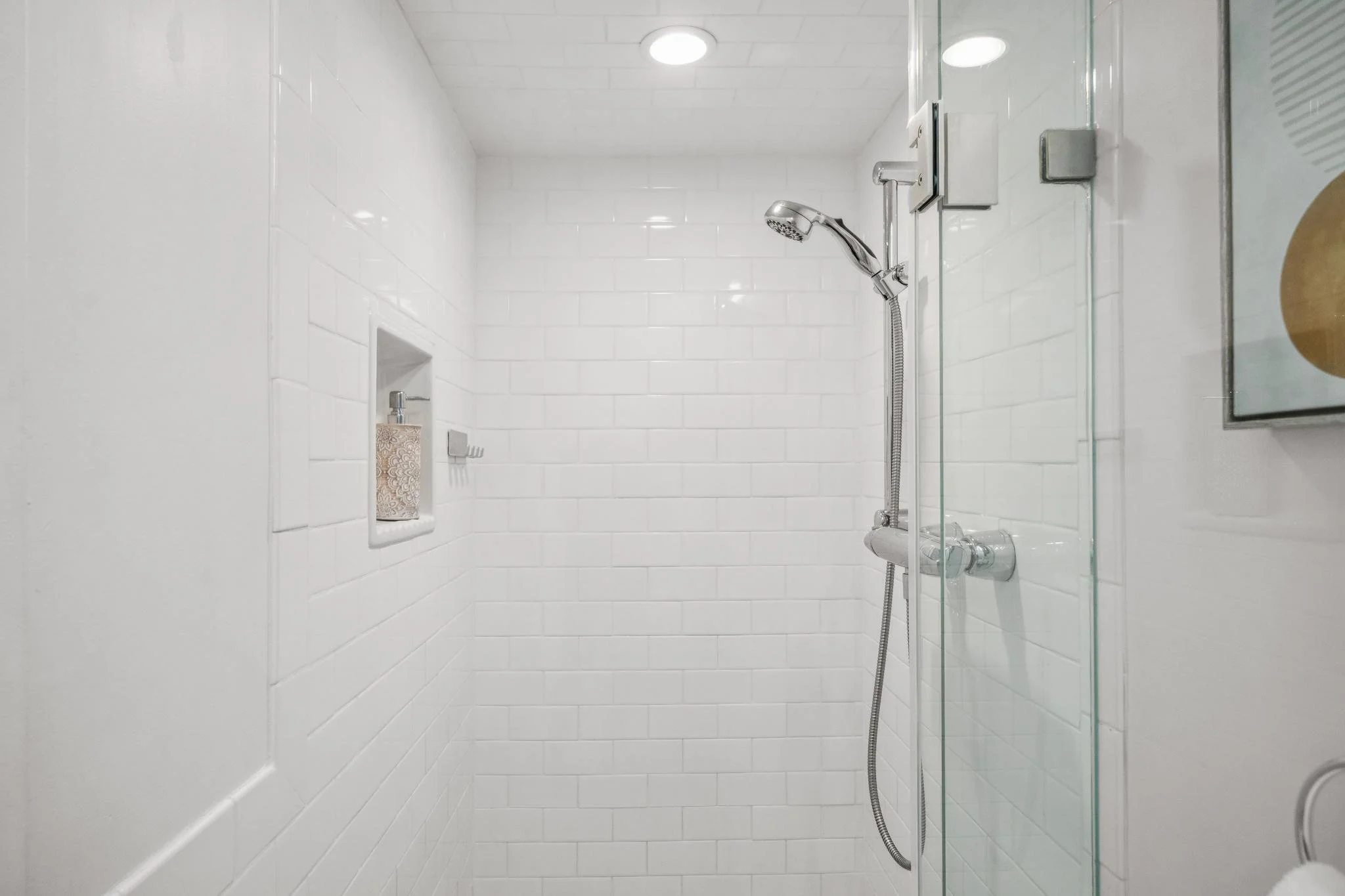 Empty shower with white tile walls, a silver showerhead, a built-in soap niche, and glass door.