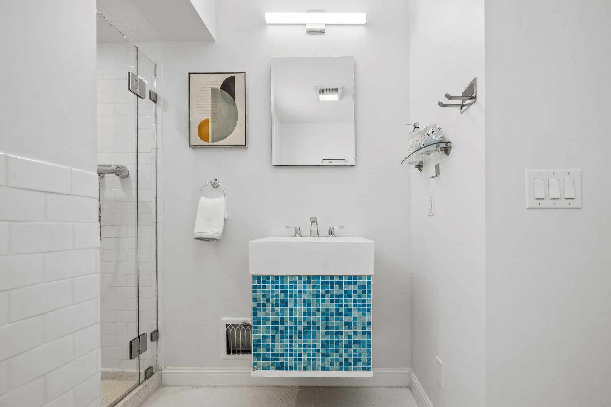 A modern bathroom with white walls, a small white sink with blue mosaic tiles on the front, a rectangular mirror, wall art, a towel holder with a white towel, and a glass shelf holding decorative items. There is a shower with a glass door on the left