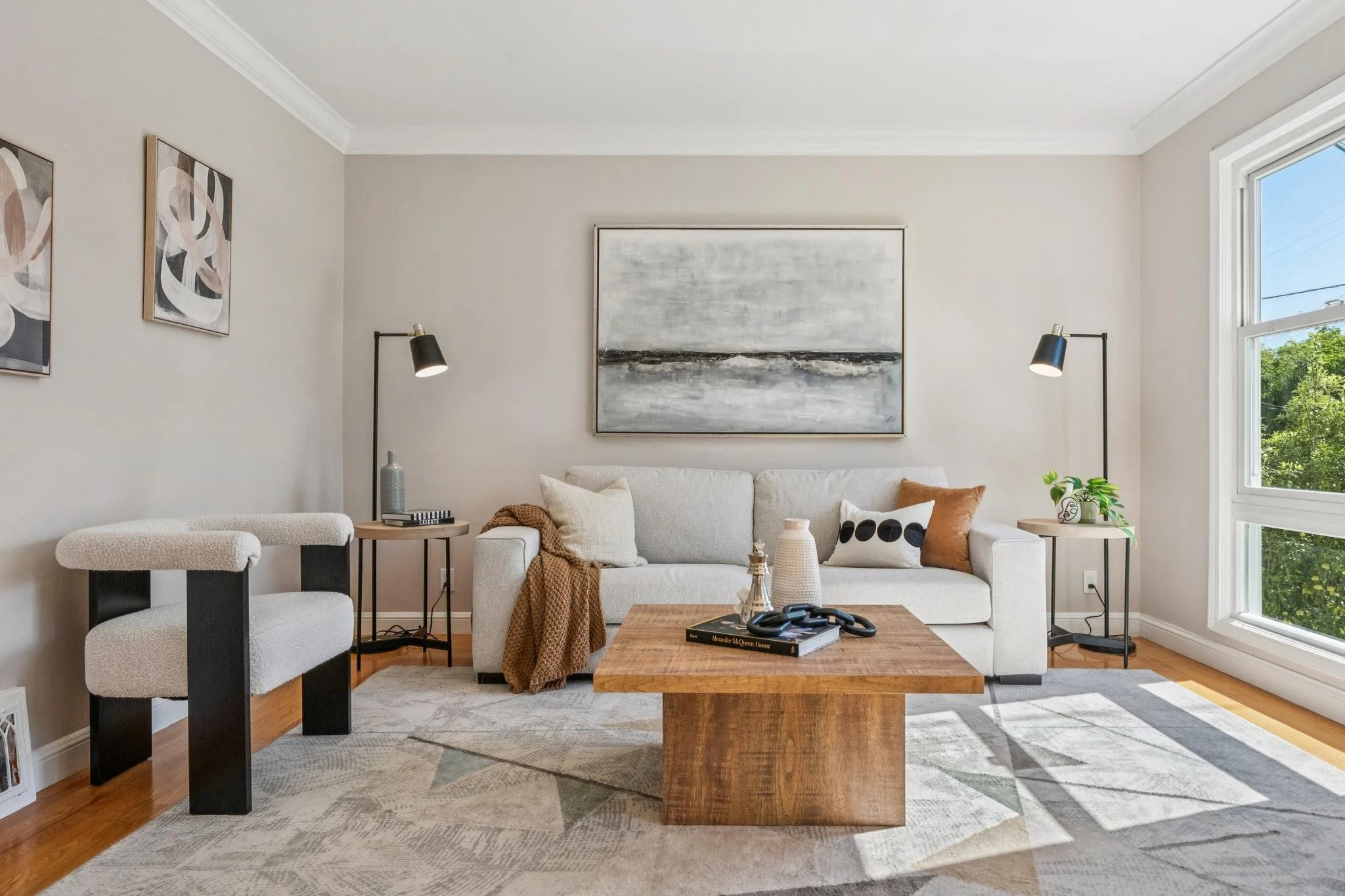 Living room with a white sofa, wooden coffee table, abstract artwork on the wall, side tables with lamps, plants, and a large window showing trees outside.