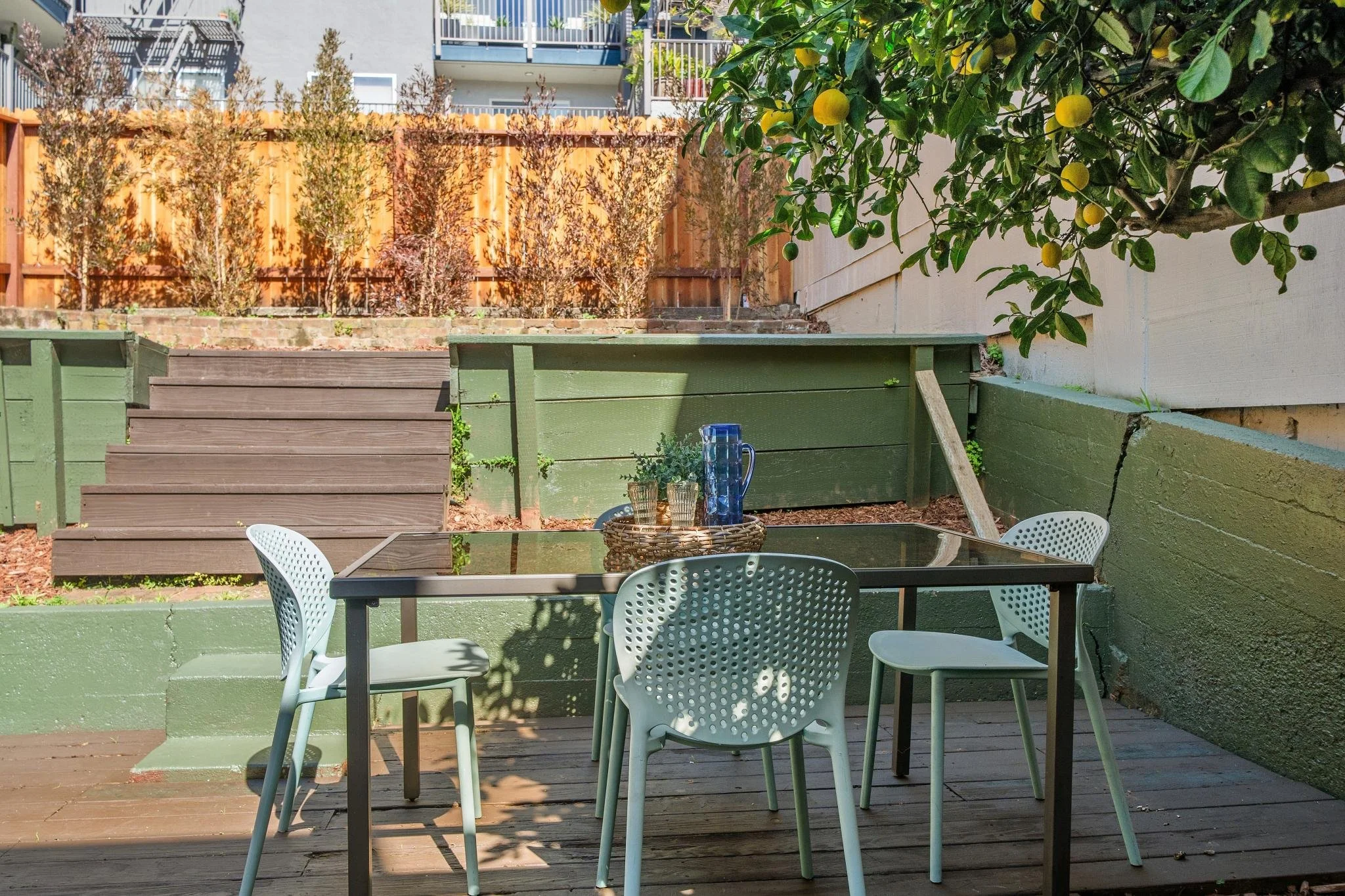 A backyard patio with a black table, four white modern chairs, a tray with glasses and a pitcher, wooden stairs leading to raised garden beds, a citrus tree with yellow fruit, and a wooden fence with small bushes behind it.