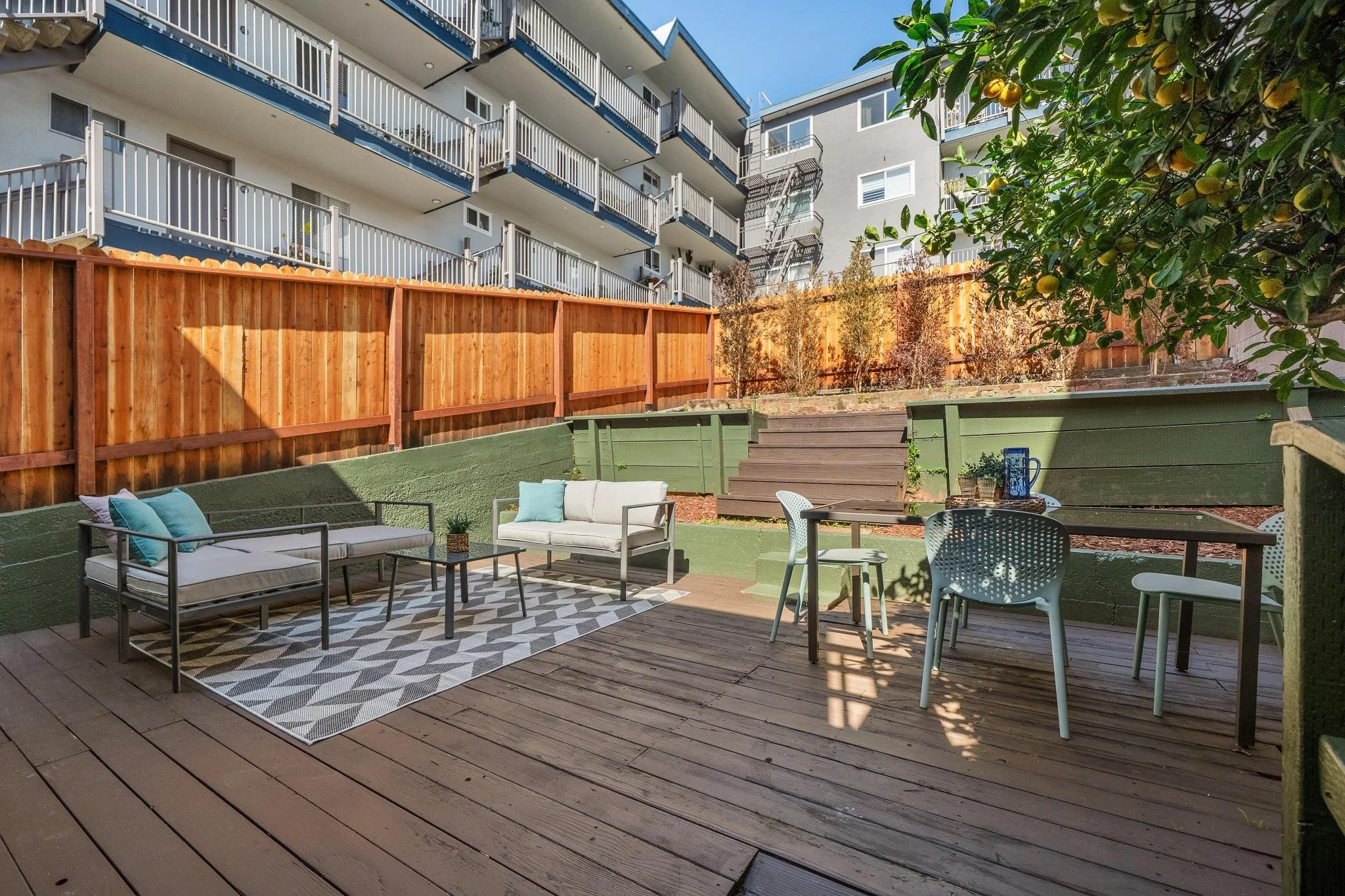 Backyard patio area with outdoor furniture, including a white cushioned sofa, a matching white cushioned loveseat, and a patio table with four chairs. There is a patterned outdoor rug, potted plants, and a wooden fence. In the background, there are s