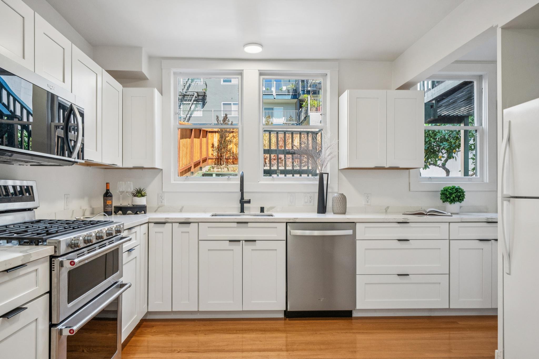 Modern white kitchen with stainless steel appliances, wooden flooring, and large windows overlooking a backyard with a wooden fence and neighboring buildings.