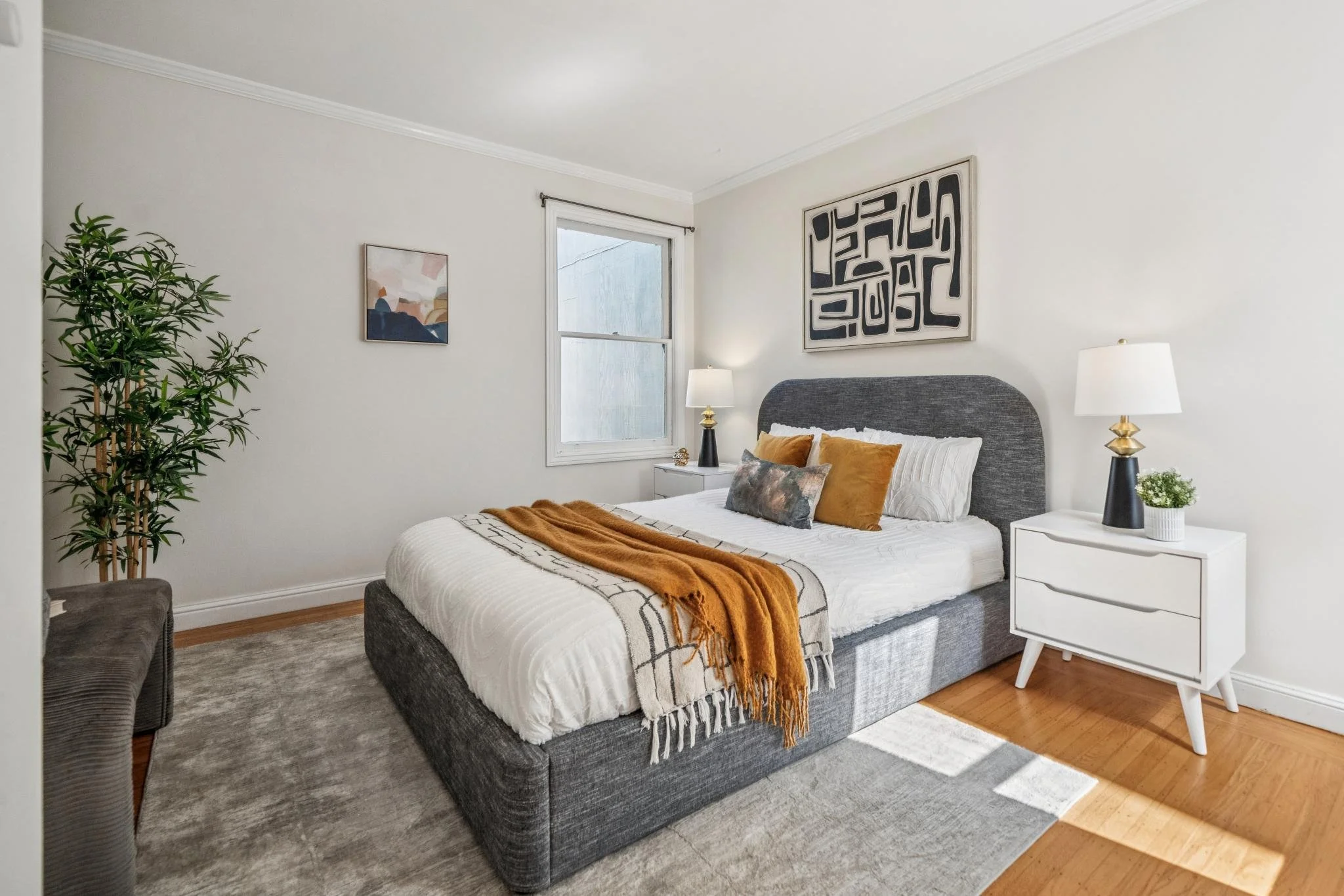 A modern bedroom with a gray upholstered bed, white bedding, and orange, gray, and white pillows. There are two white nightstands with black and gold lamps, a potted plant, a cat-themed artwork, and an abstract wall art. A large window lets in natura