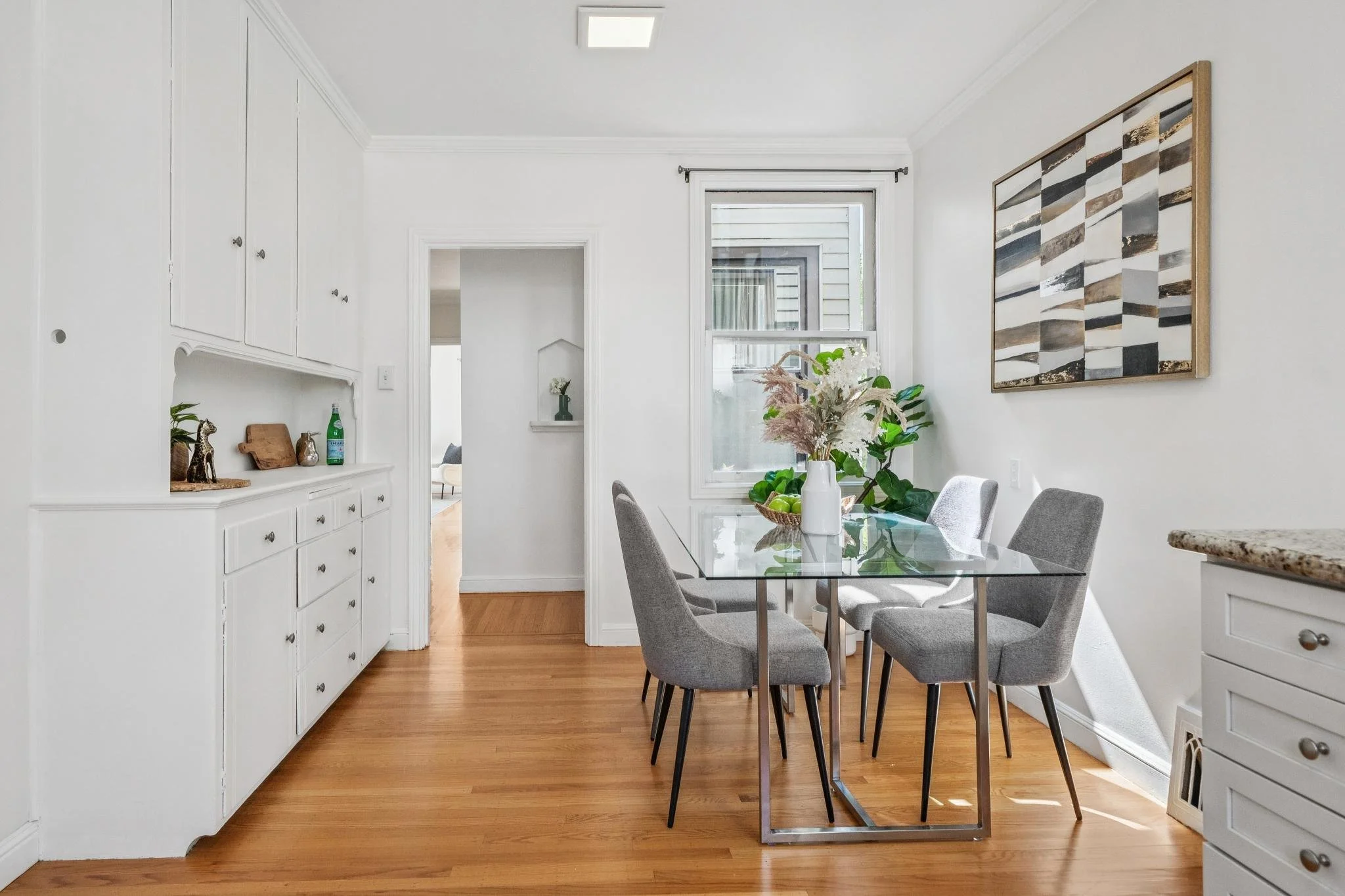 A dining room with a glass table and six light gray upholstered chairs, a white vase with flowers and green apples on the table, a window with outside view, white cabinets, and a colorful abstract wall art.