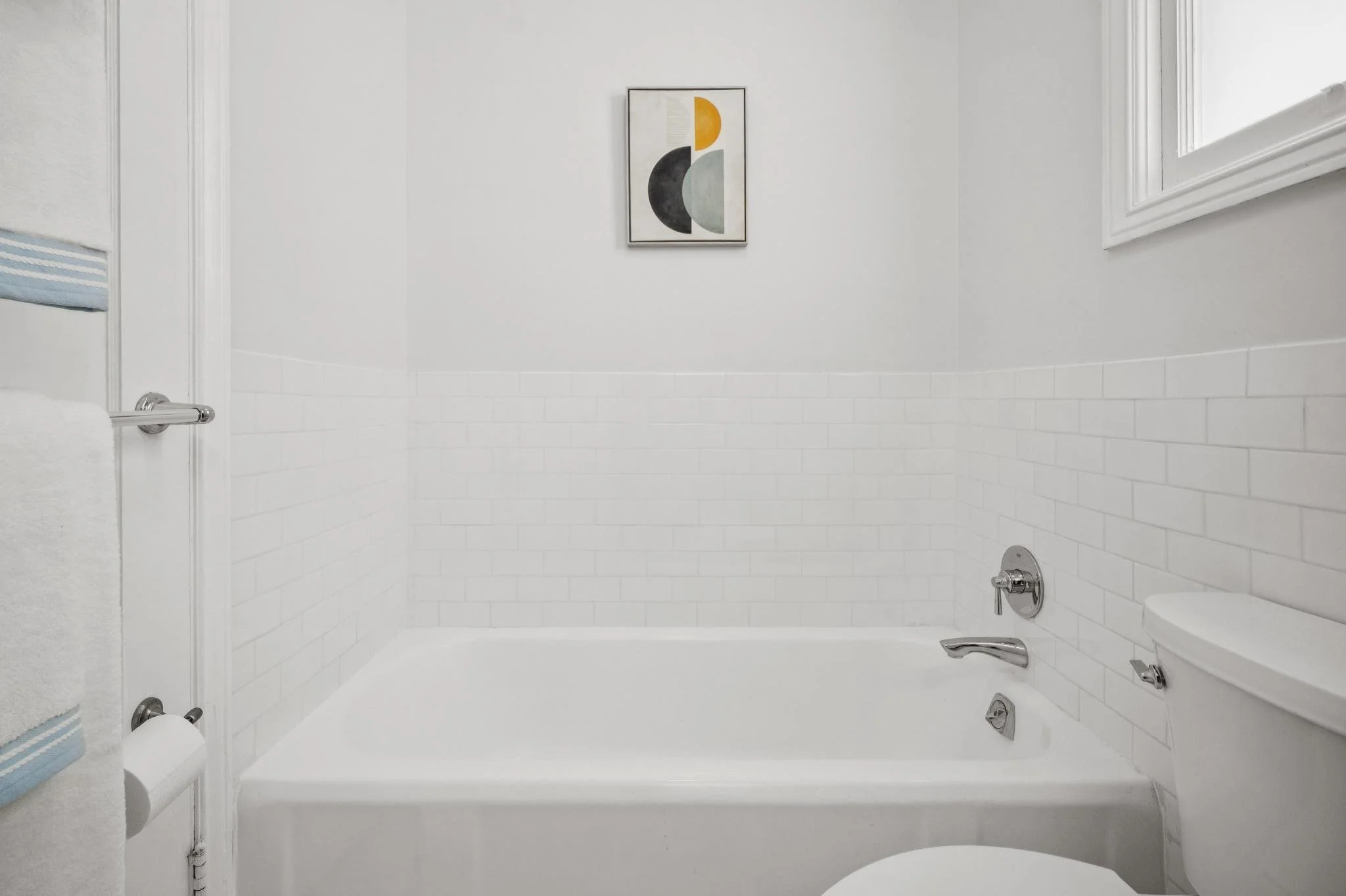 Bathroom with a bathtub, white tile walls, a framed abstract art piece, a window, and a towel on a towel rack.