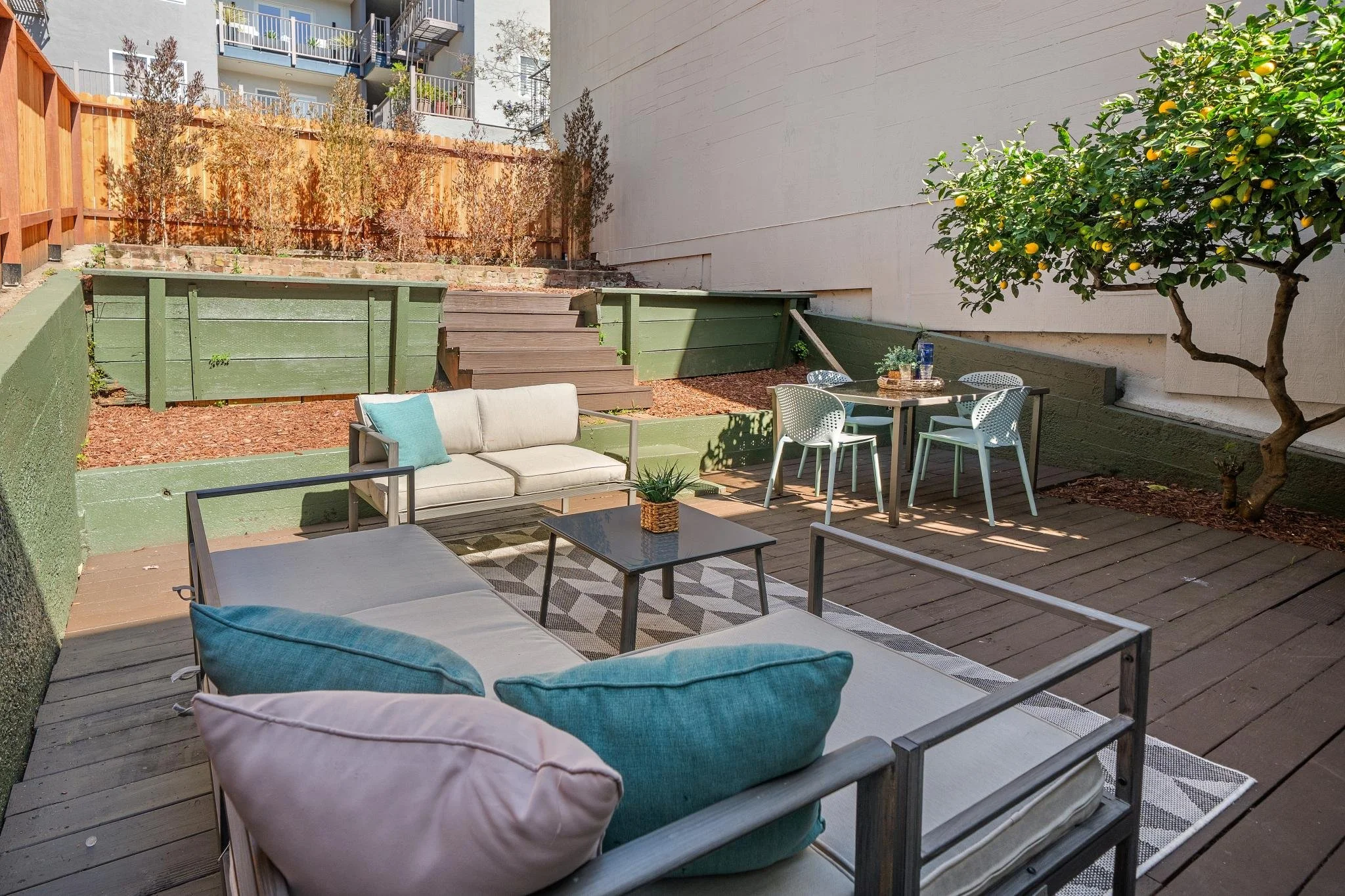 Outdoor patio with seating area, including a sofa, chairs, and tables, surrounded by a wooden deck and planting beds, with a lemon tree on the right.
