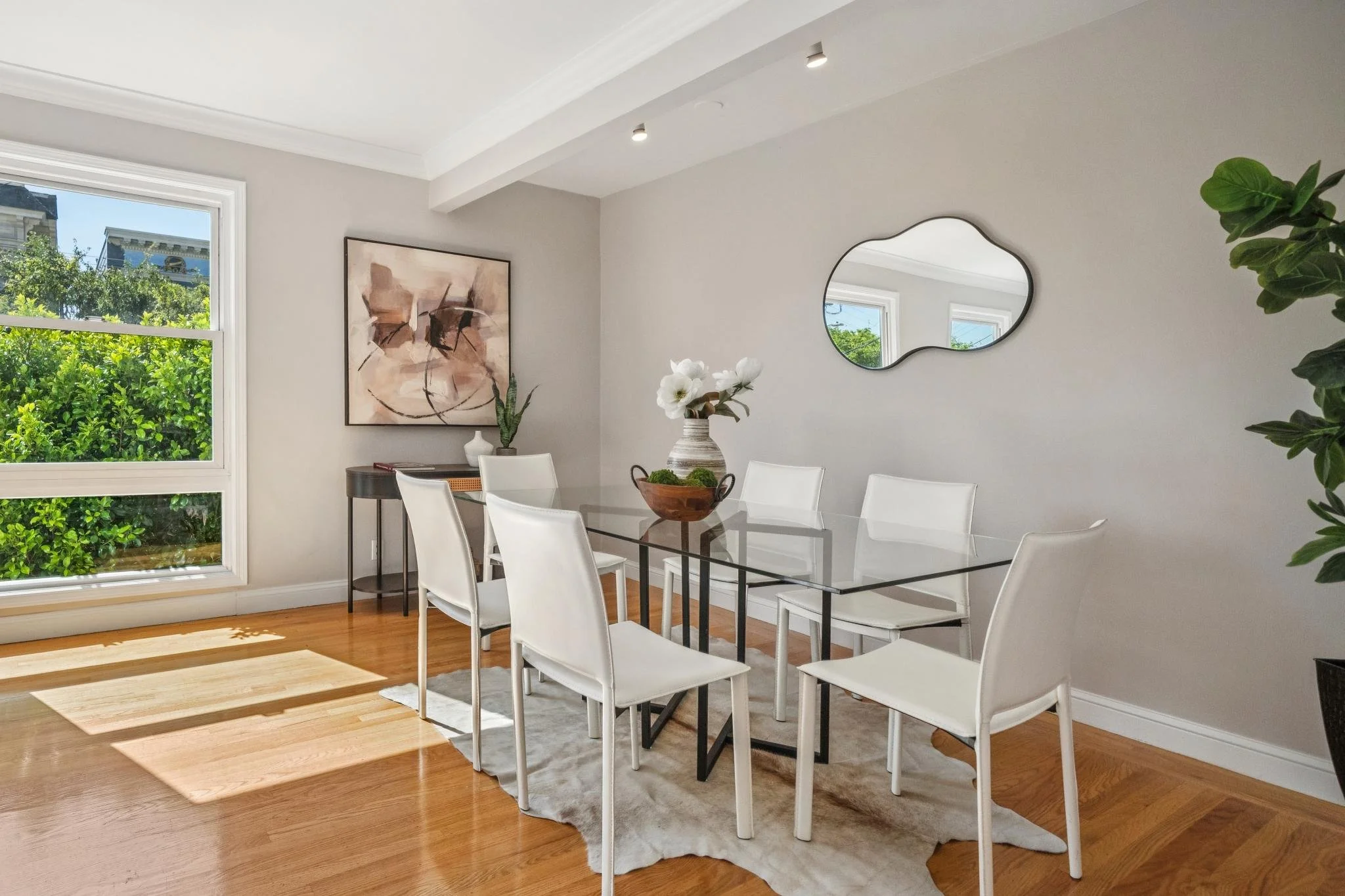 Dining room with a glass table, six white chairs, a wooden bowl with flowers, artwork on the wall, a mirror, and hardwood floors.