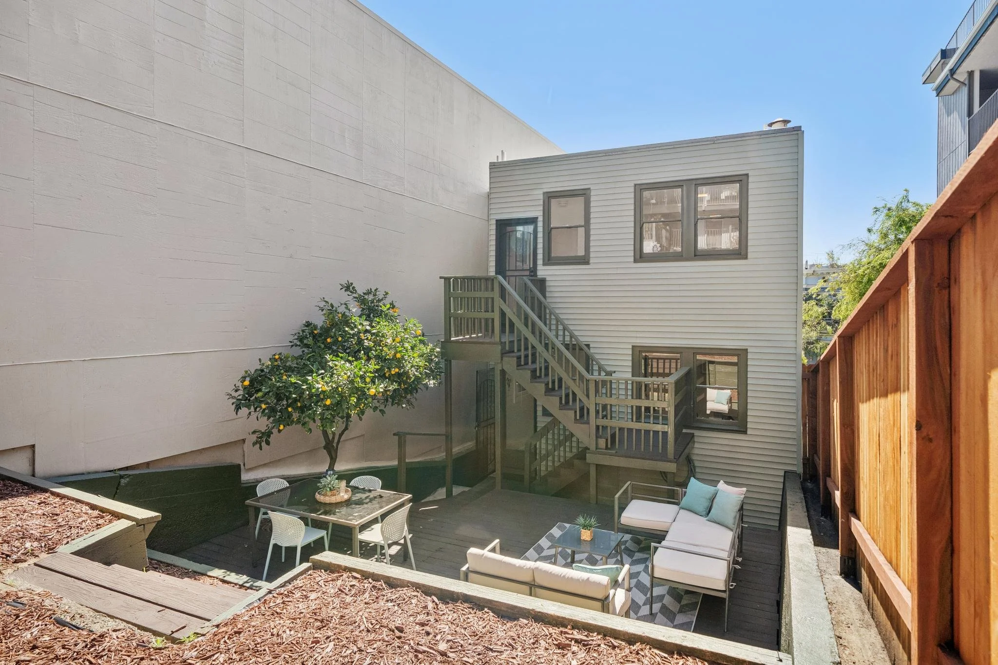 Small backyard patio with outdoor furniture, including a sofa, armchairs, coffee table, and a dining table with chairs, surrounded by a wooden fence and adjacent to a two-story house with gray siding and multiple windows, with a lemon tree and mulch 