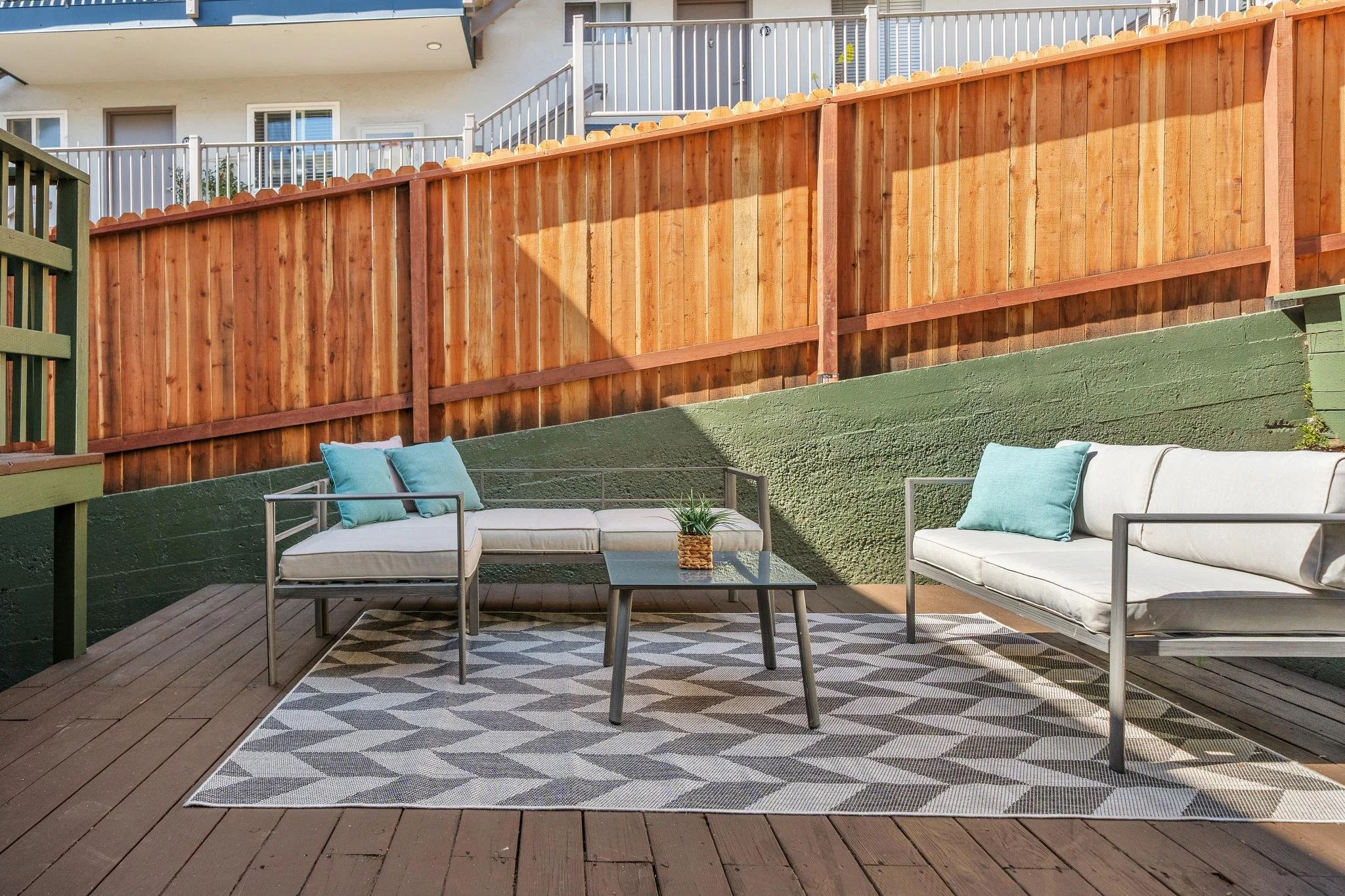 Outdoor patio with two white cushioned sofas with metal frames, teal pillows, a small black coffee table with a potted plant, a patterned outdoor rug, wooden deck, and a wooden fence with a house in the background.