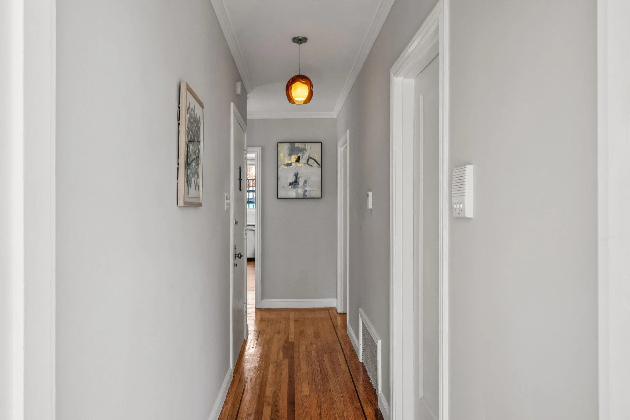 Empty hallway with light gray walls, a wooden floor, and a ceiling light fixture. There are framed artworks on the walls and a door at the end of the hall.