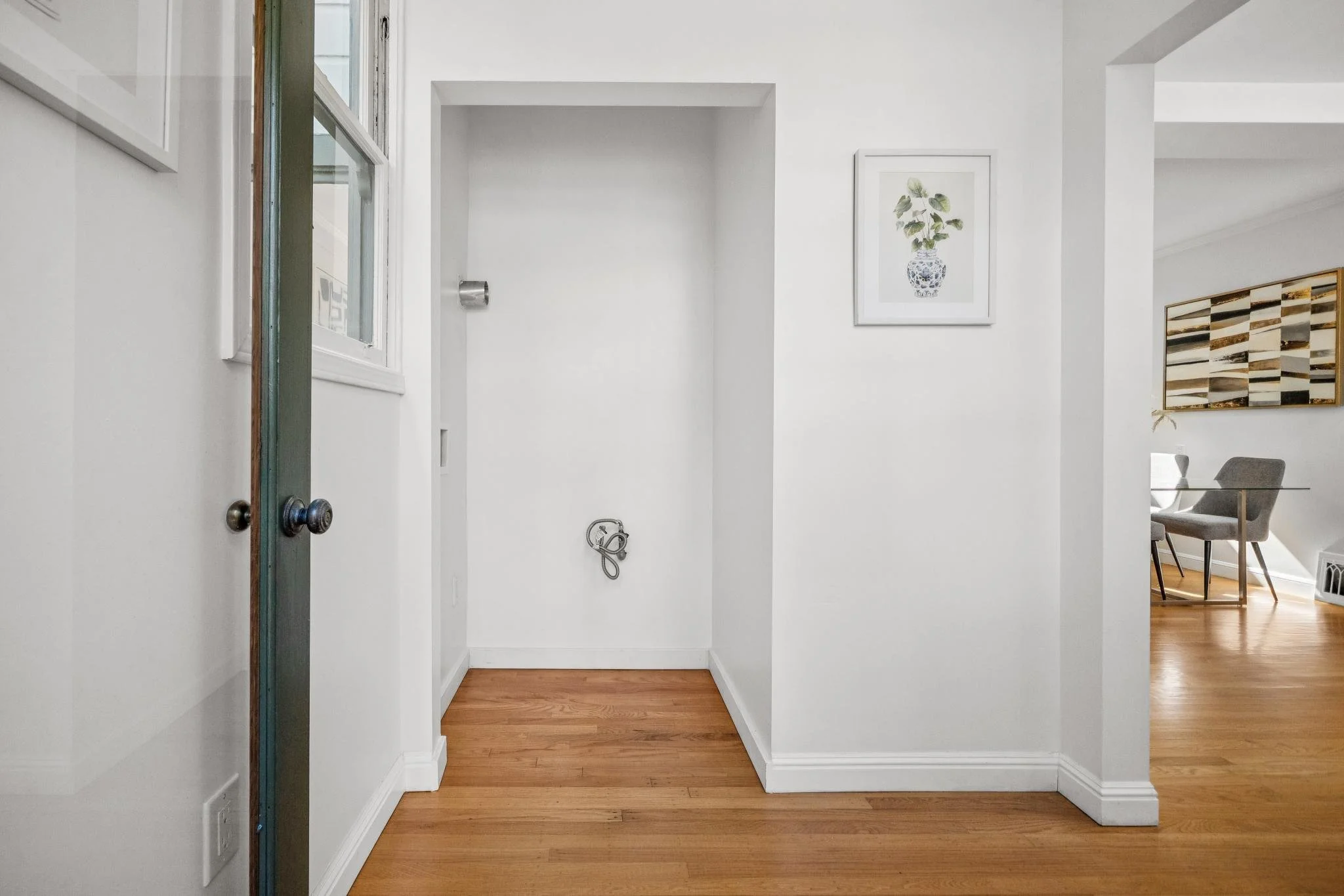 Empty white wall space in a house with a small alcove for a washing machine, with hookups visible, wooden flooring, and a sliding glass door to the left.