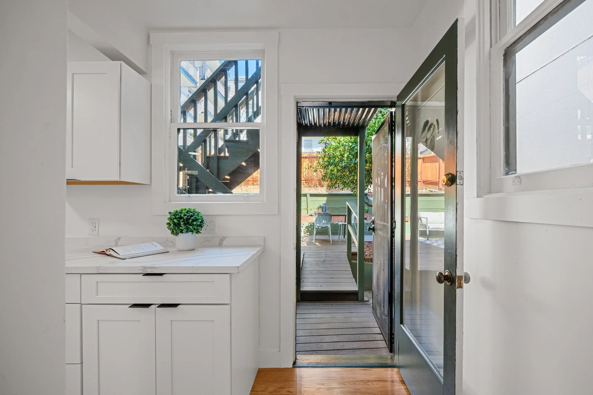 Interior view of a kitchen with white cabinets and marble countertop, a small potted plant, and an open door leading to a backyard patio with chairs and a green fence.