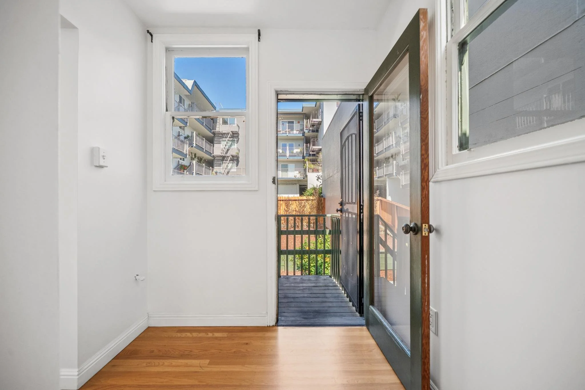 View from inside an apartment looking toward the front door with a screen door open, showing a small balcony and neighboring apartment buildings outside.