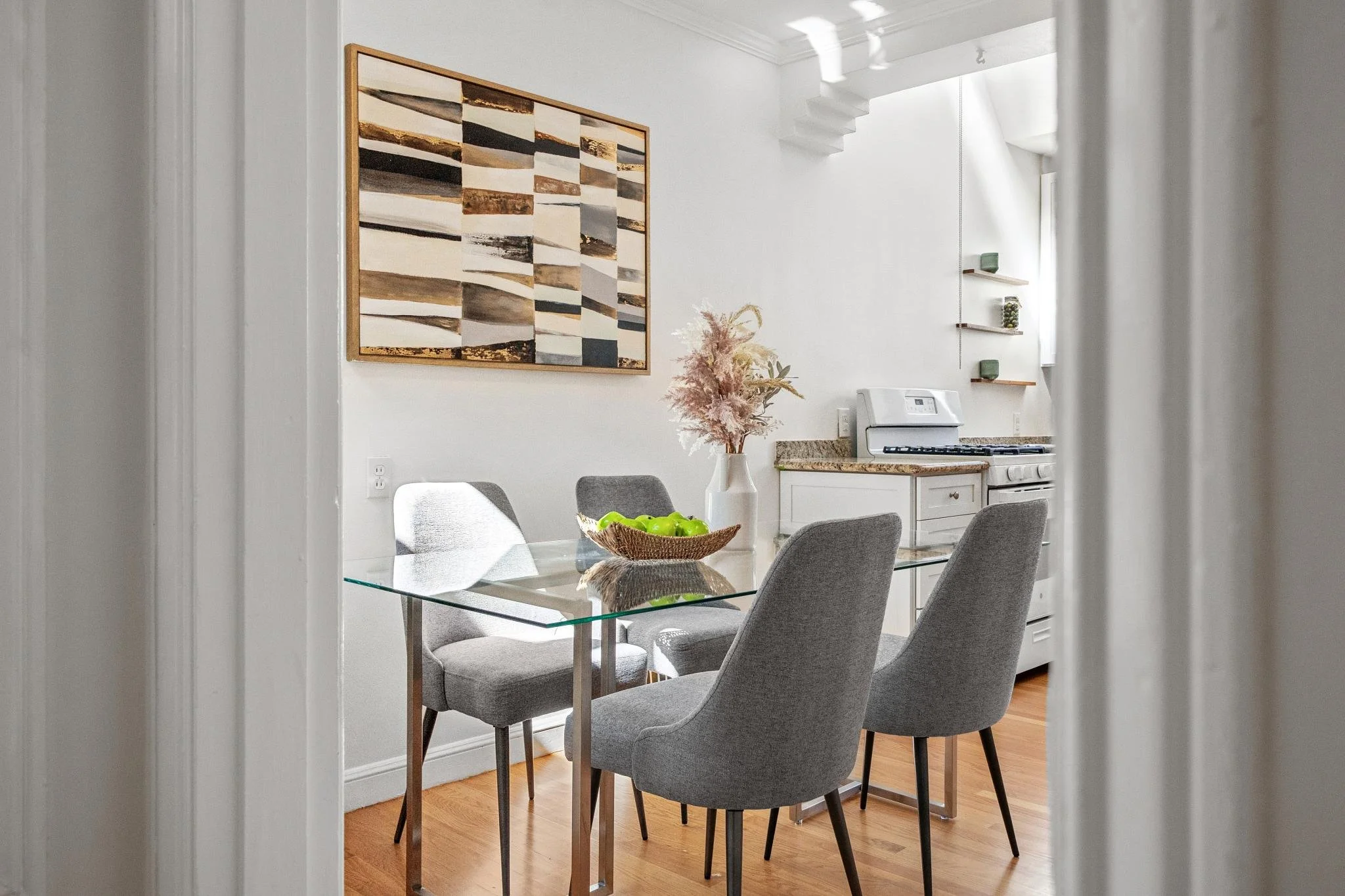 Modern dining room with a glass table, four grey fabric chairs, a vase with pink pampas grass, a wooden bowl with green apples, and a large abstract painting on white walls. Part of the kitchen with white cabinets, granite countertops, and a stove is