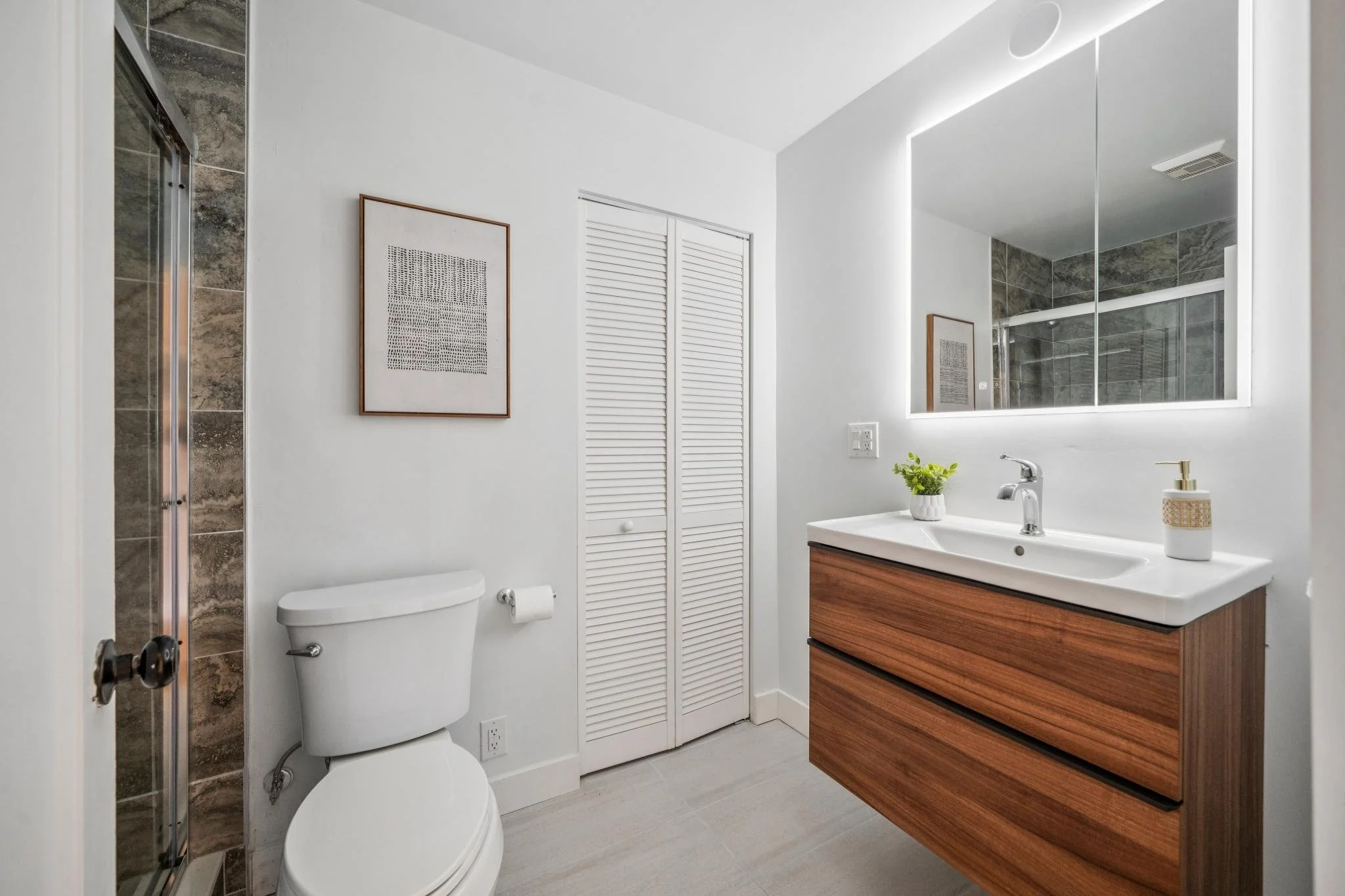 Modern bathroom with white walls, wooden vanity, large mirror with LED lighting, small potted plant, soap dispenser, toilet, and a shower with a glass door and ceramic tile walls.