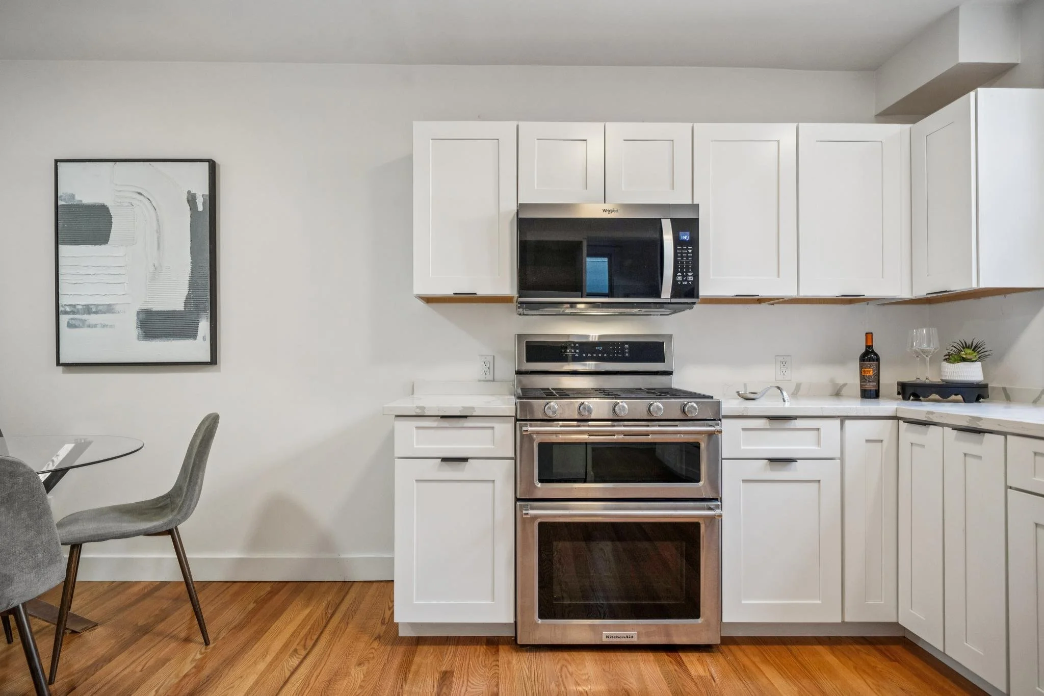 Kitchen with white cabinets, stainless steel microwave, oven, and stovetop, white countertops, wine bottle, wine glass, plant, and abstract wall art.