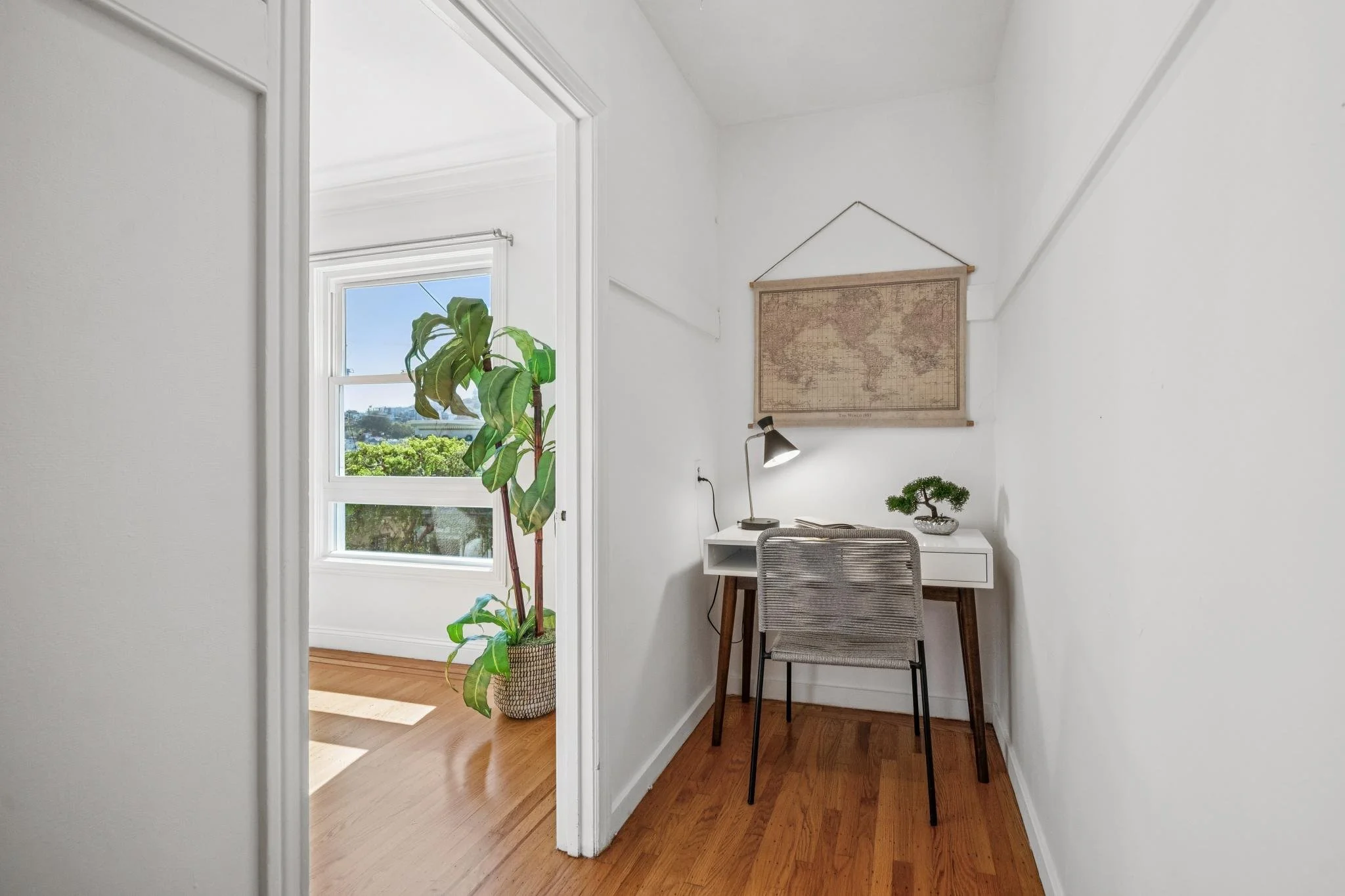 Small home office nook with white walls, a white desk with wooden legs, a gray chair, a small desk lamp, a tucked away window with a large green plant nearby, and a wall hanging map.