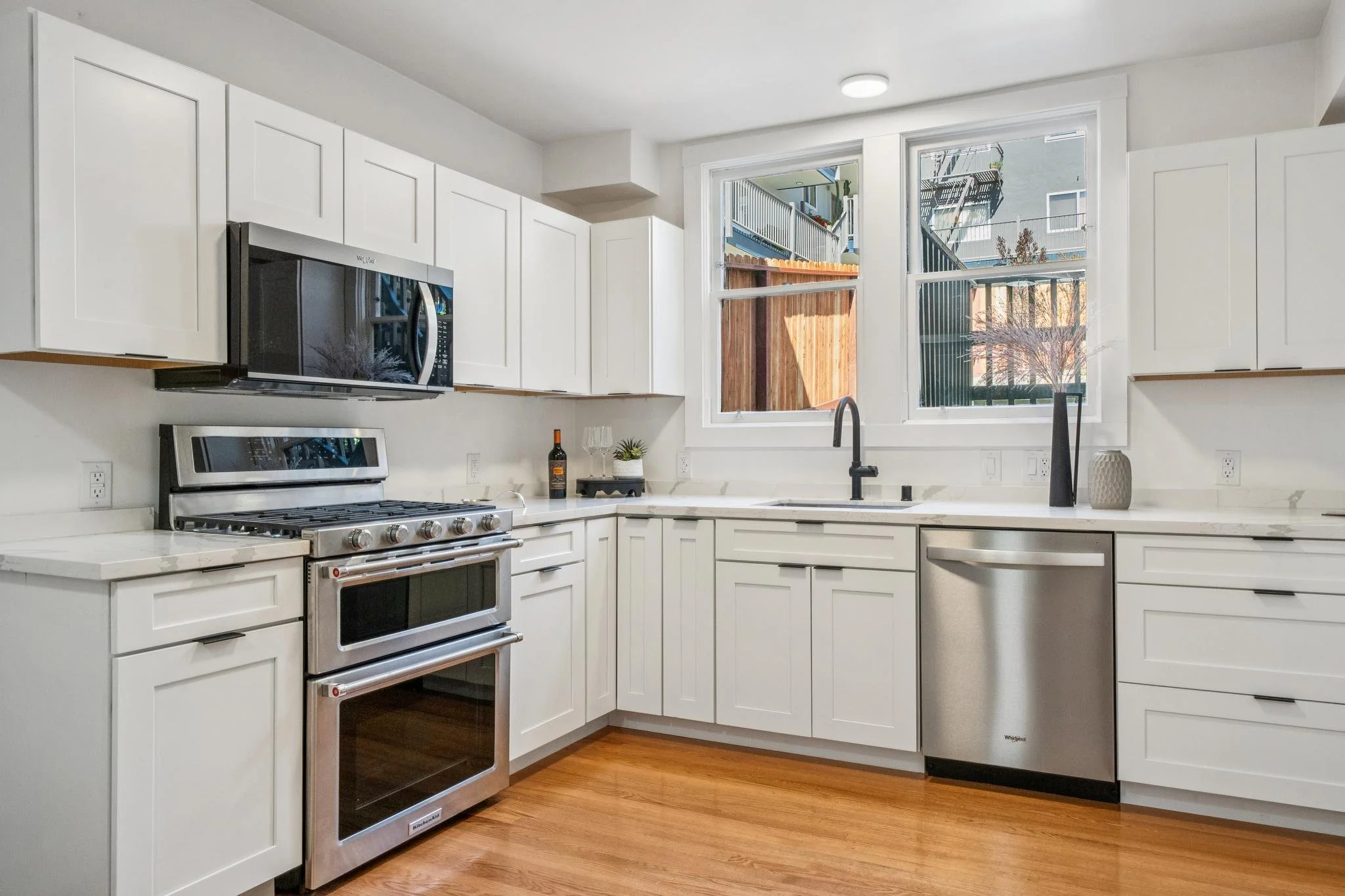 Modern white kitchen with stainless steel appliances, black faucet, and large windows showing outdoor patio area.
