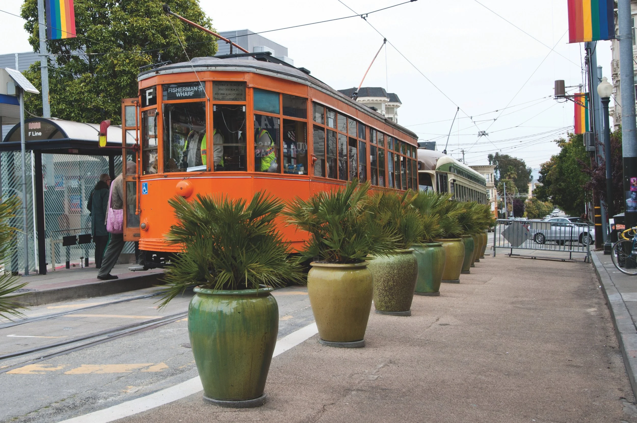 Orange historic streetcar at a station with people boarding, lined with large potted plants, on a city street with trees and buildings in the background.