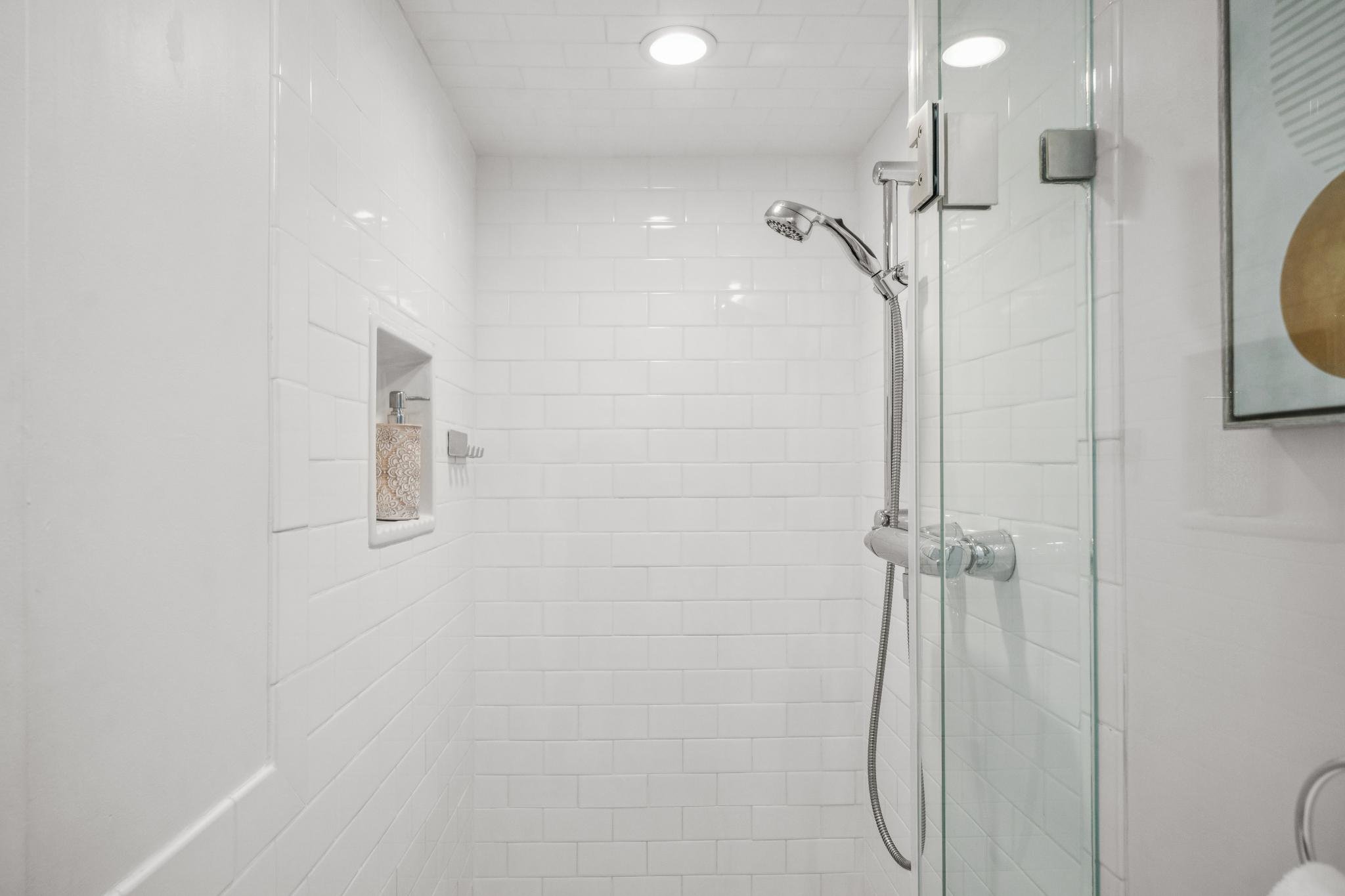 A clean, white tiled shower with a glass door, handheld showerhead, and built-in soap niche.