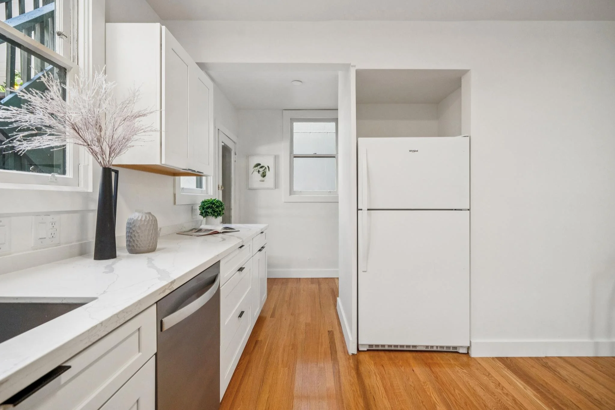 A modern kitchen with white cabinets, a white marble countertop, a white refrigerator, and wooden flooring. There are decorative vases and a plant on the counter and two windows letting in natural light.