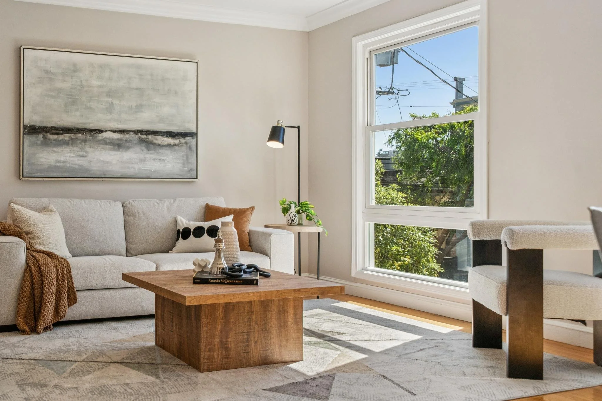 Bright living room with a white sofa, abstract black and gray painting, wooden coffee table with decor, small side table with green plant, floor lamp, large window with greenery outside, and a white upholstered chair.