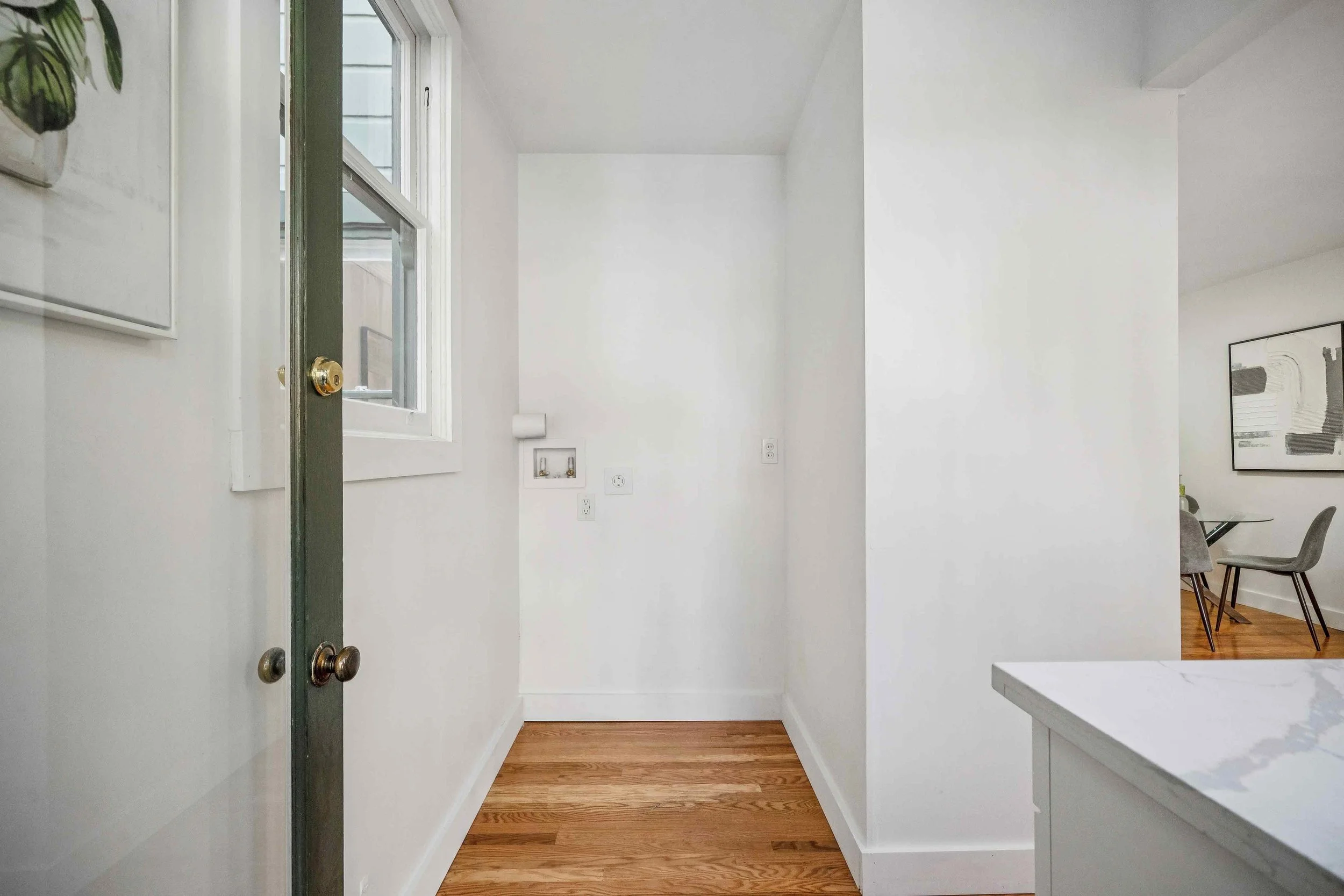 Empty white kitchen corner with hardwood floor, window, and part of dining area with chairs and artwork on wall.