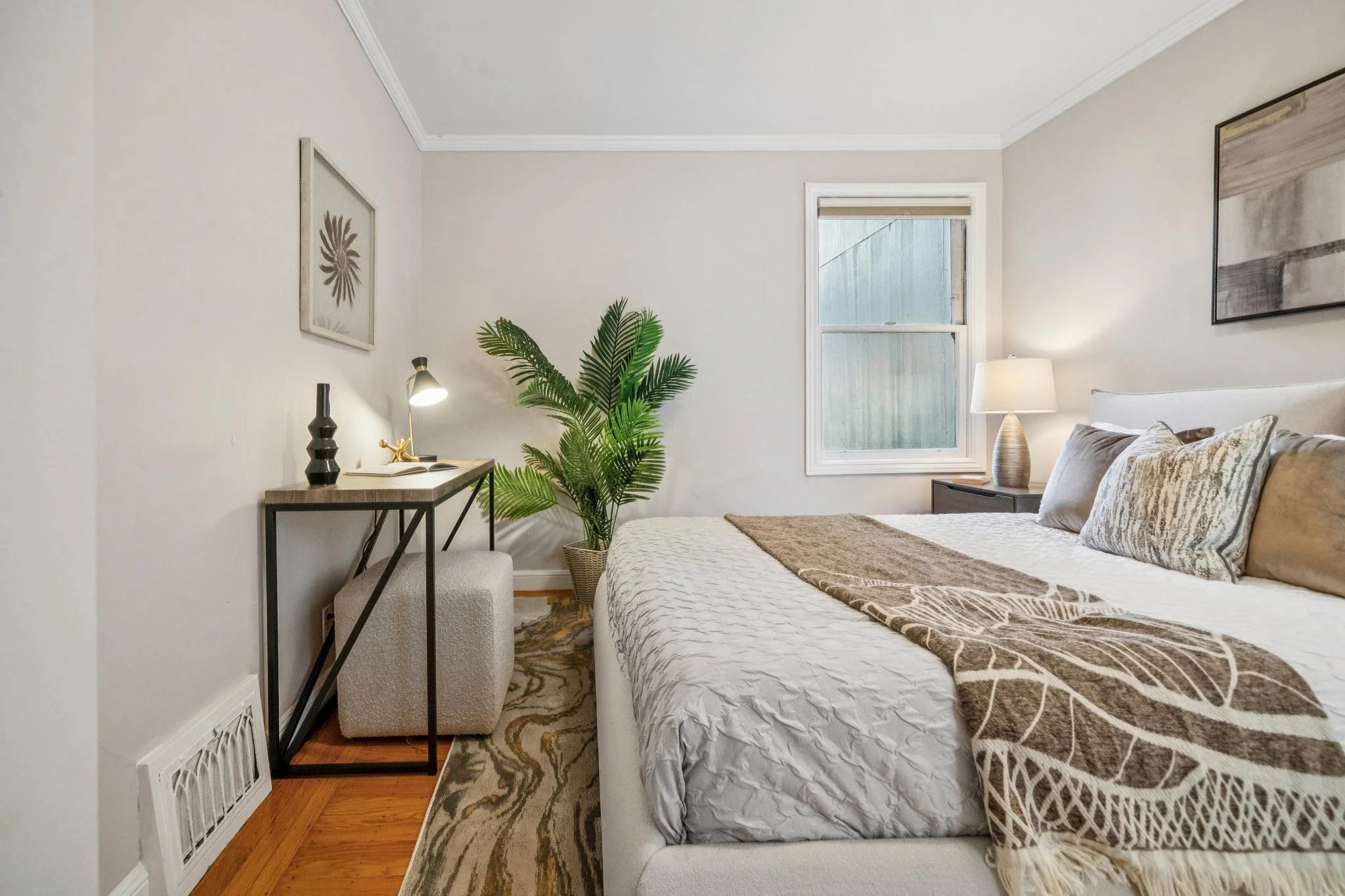 A cozy bedroom with a large bed, gray pillows, nightstand with a lamp, a potted plant, a window, and a small desk with a lamp and decorative items.