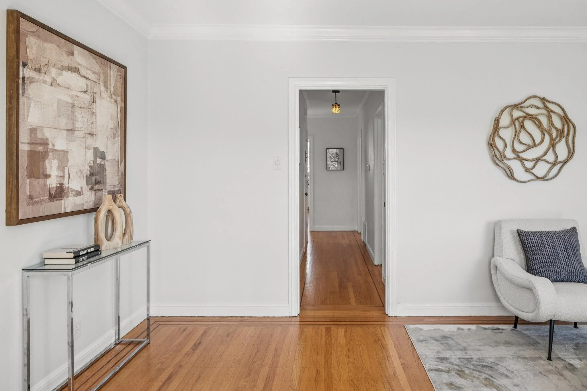 Living room with white walls and hardwood floors, door leading to hallway with additional artwork, decorative wall art, modern chair with pillow, glass top console with books and sculptures, and abstract wall art.