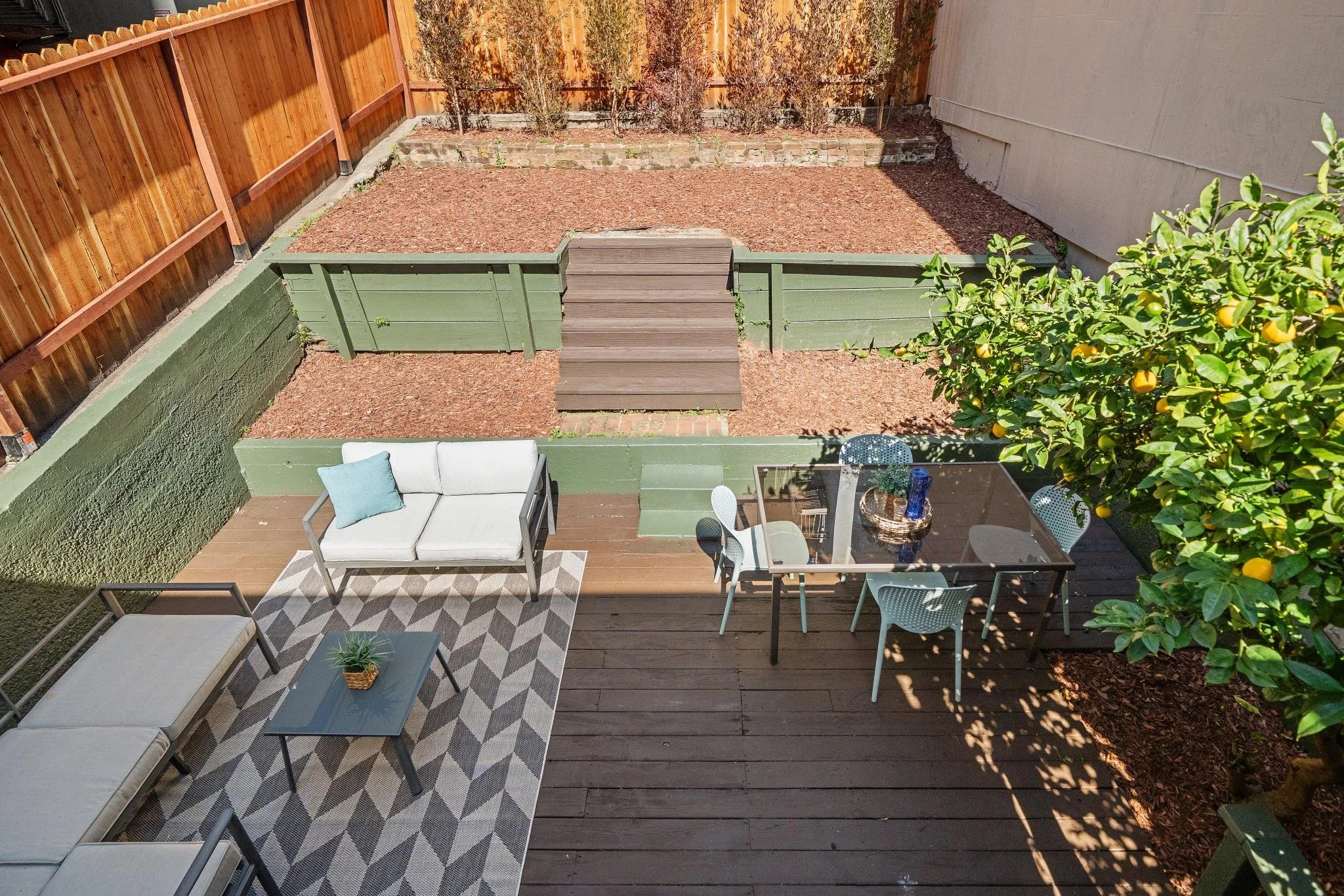 An outdoor backyard patio with seating area, including a white cushioned outdoor sofa with a blue pillow, a small black coffee table with a plant, and a glass dining table with four chairs. The yard is divided into two sections: a deck area and a gro