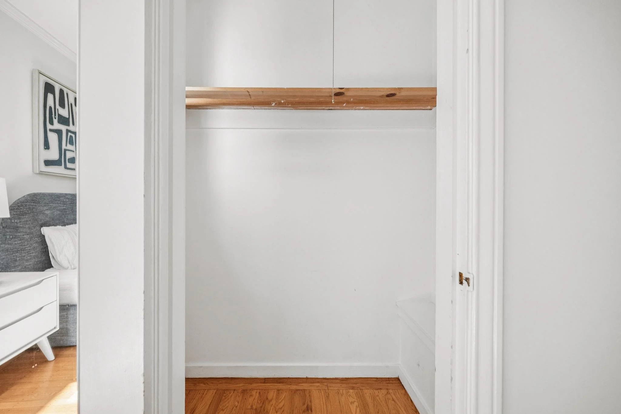 Empty closet with a wooden shelf and white walls, part of a bedroom with a bed and artwork.