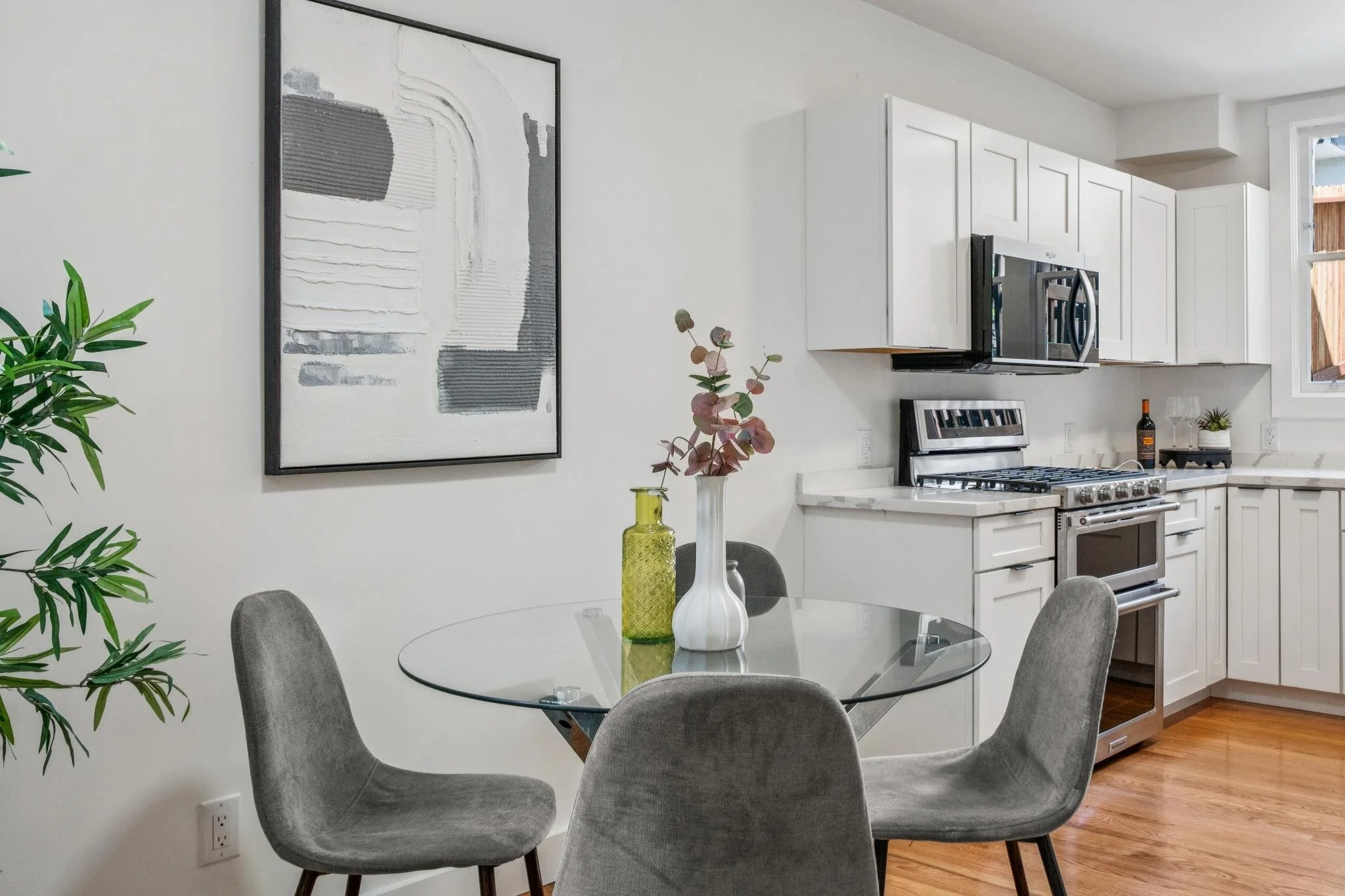Modern kitchen with white cabinets, stainless steel stove and microwave, round glass dining table with gray upholstered chairs, decorative vases, abstract wall art, and hardwood flooring.