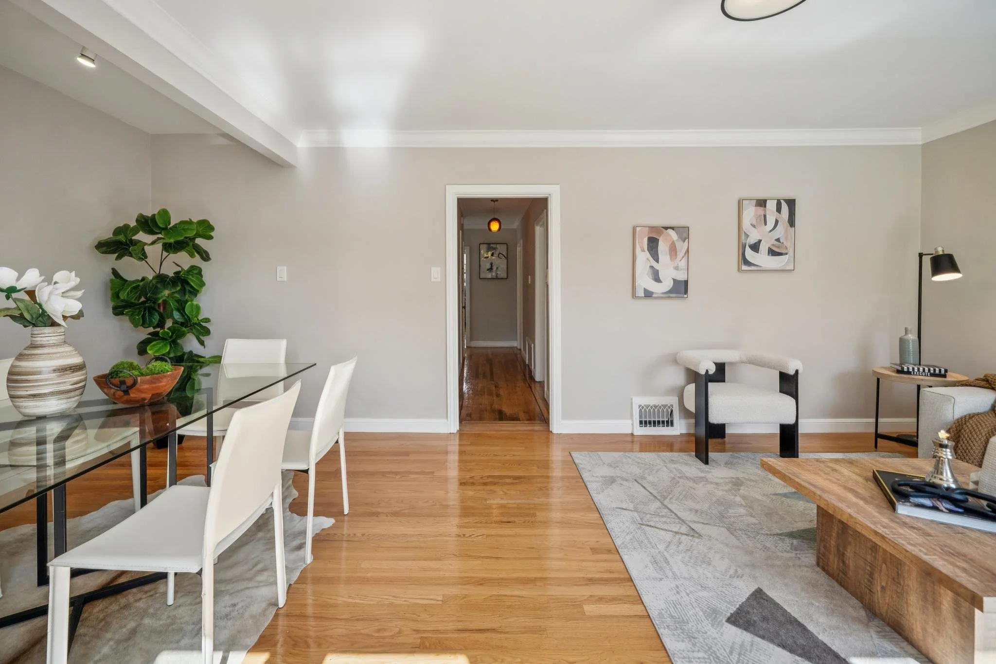 Living room with hardwood floors, light-colored walls, modern artwork, a glass dining table with white chairs, a white armchair, a black floor lamp, a beige sofa with a wooden coffee table, and decorative items including vases and artwork.