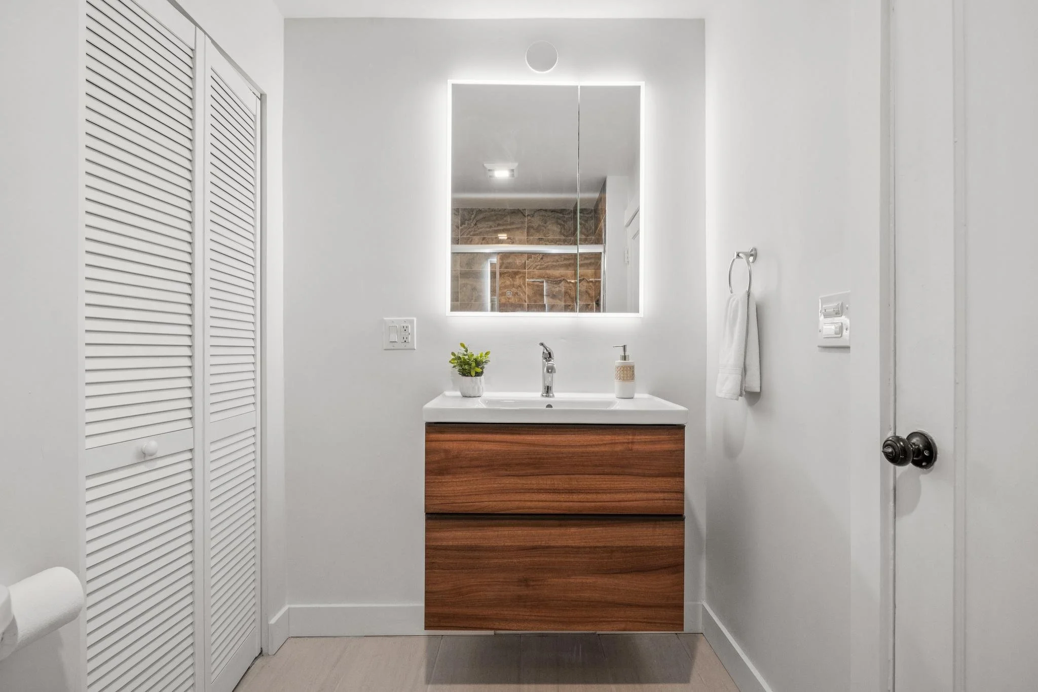 Modern bathroom with a white wall, illuminated mirror, wooden vanity with white countertop, and a small plant and soap dispenser on the counter. There is a door with black knobs, a towel rack with a white towel, and a closet with louvered doors.