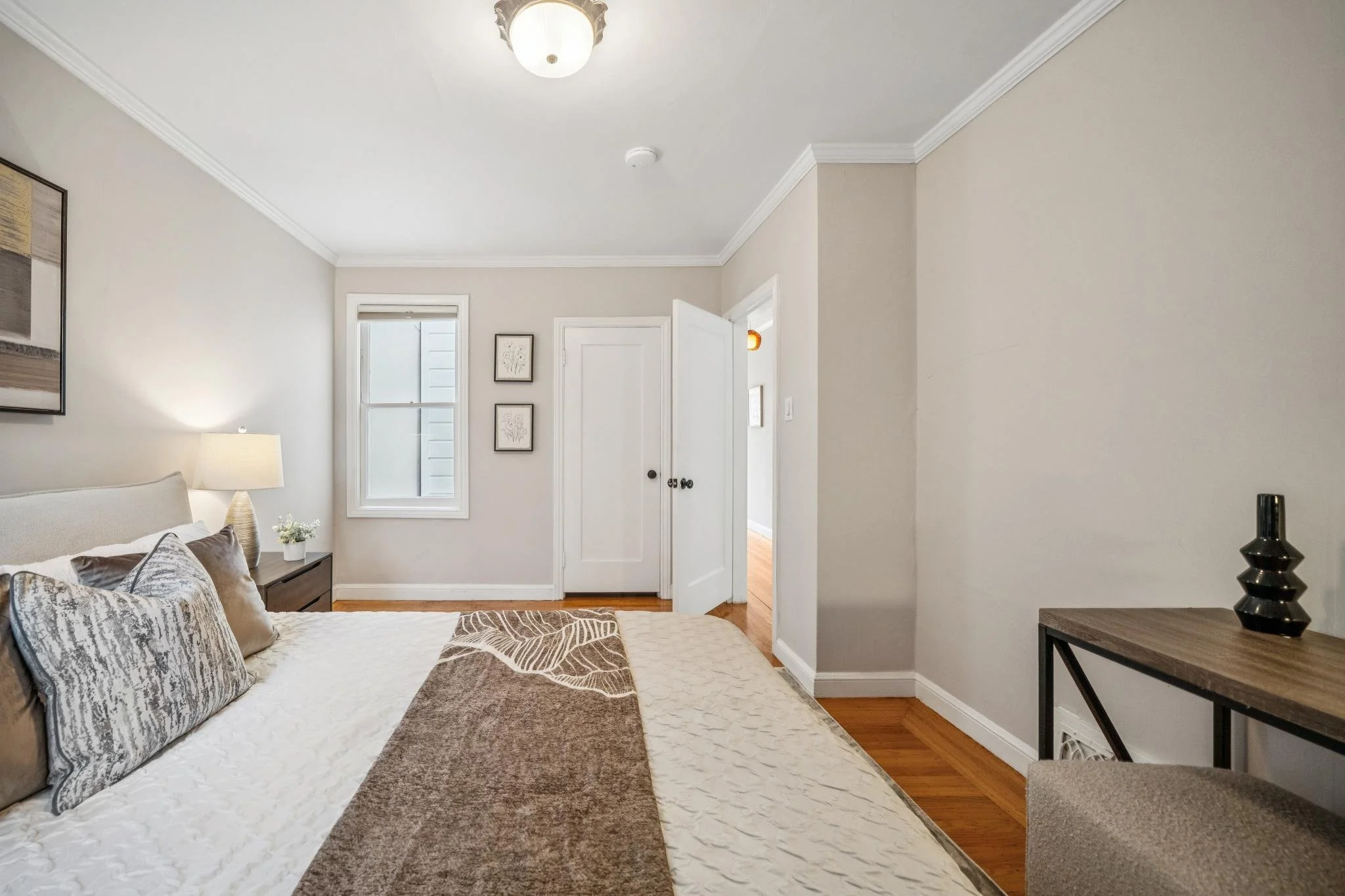 A bedroom with a bed having multiple pillows, a nightstand with a lamp, and a wall with three framed pictures. There is a window and a door in the background, with wooden flooring and minimal decor.