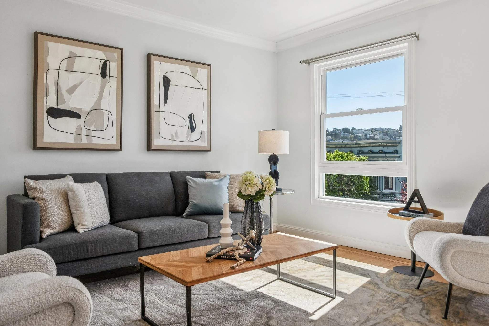 Living room with a dark gray sofa, light-colored armchair, wooden coffee table, black table lamp, wall art, window with city view, and decorative items.