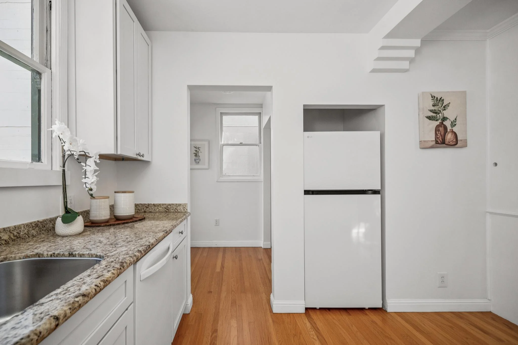 A bright kitchen with white cabinets, a granite countertop, a stainless steel sink, a white refrigerator, and hardwood floors. There are two small windows and a piece of artwork on the wall.