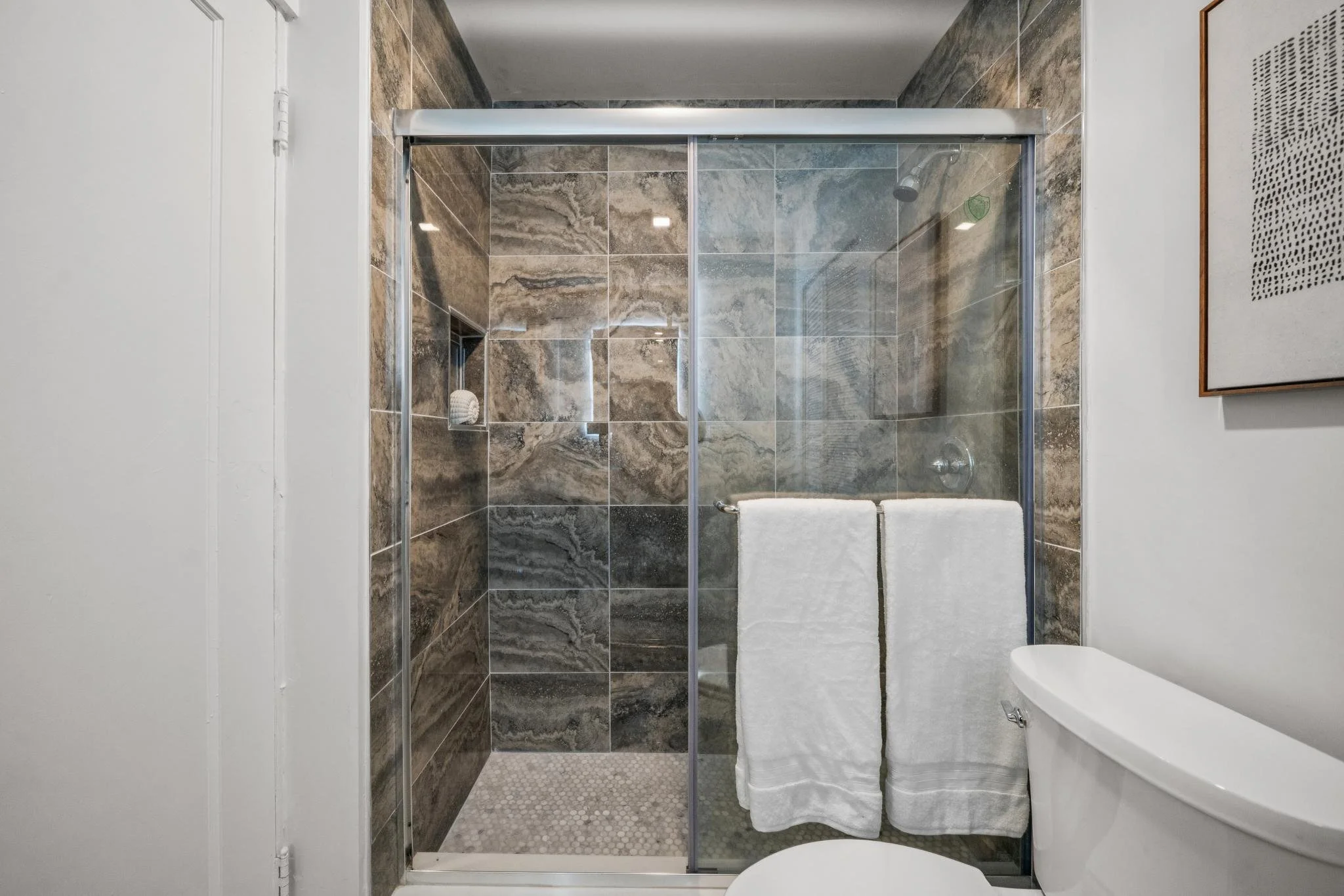 Inside a modern bathroom shower enclosure with glass doors, stone tile walls, and two white towels hanging on a towel bar. Part of a white toilet is visible in the foreground, and a framed artwork hangs on the right wall.
