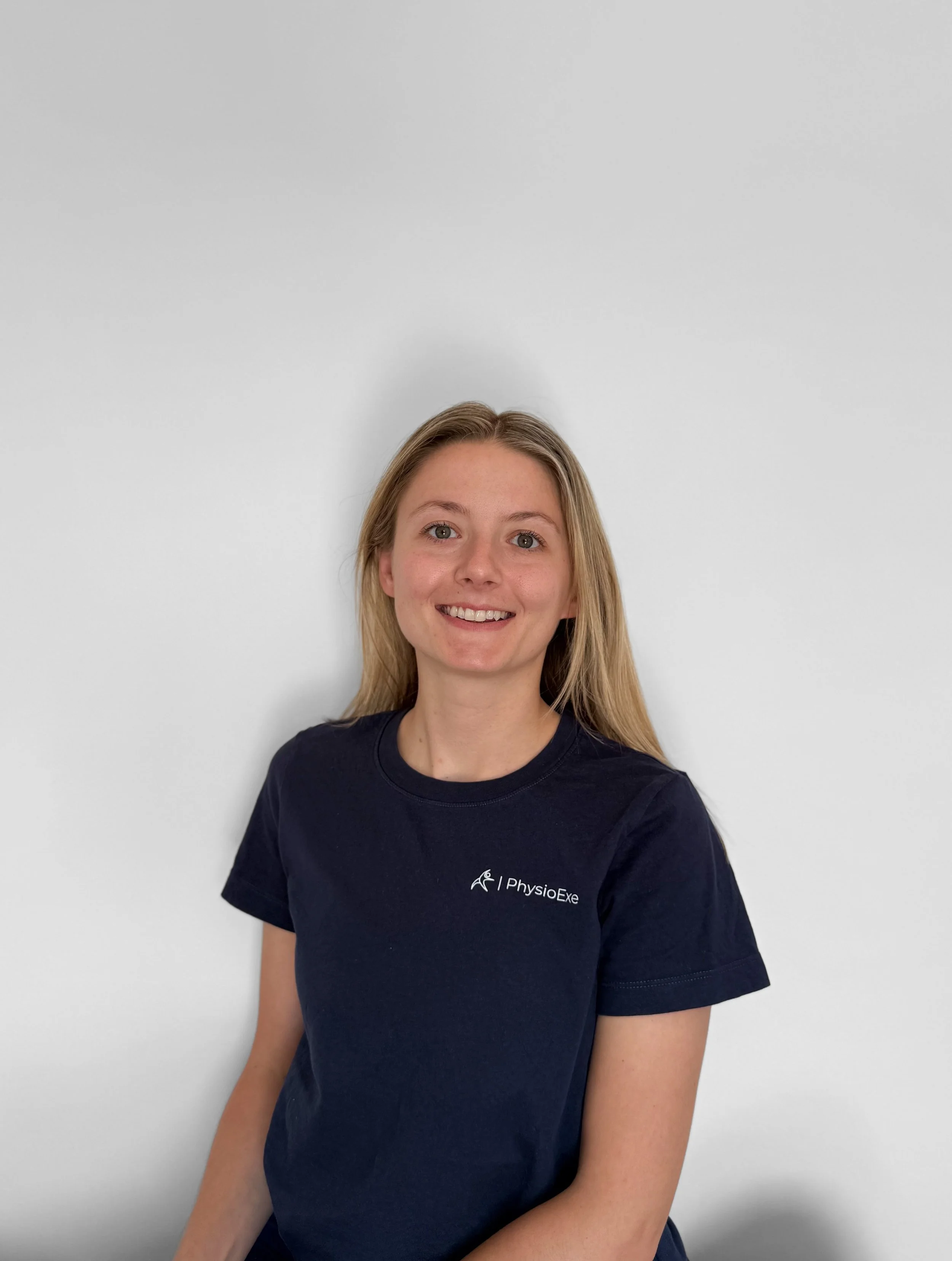 A young woman with long blonde hair smiling at the camera, wearing a navy blue T-shirt with a logo and the words 'PhysioExe' on it, standing against a plain white wall.