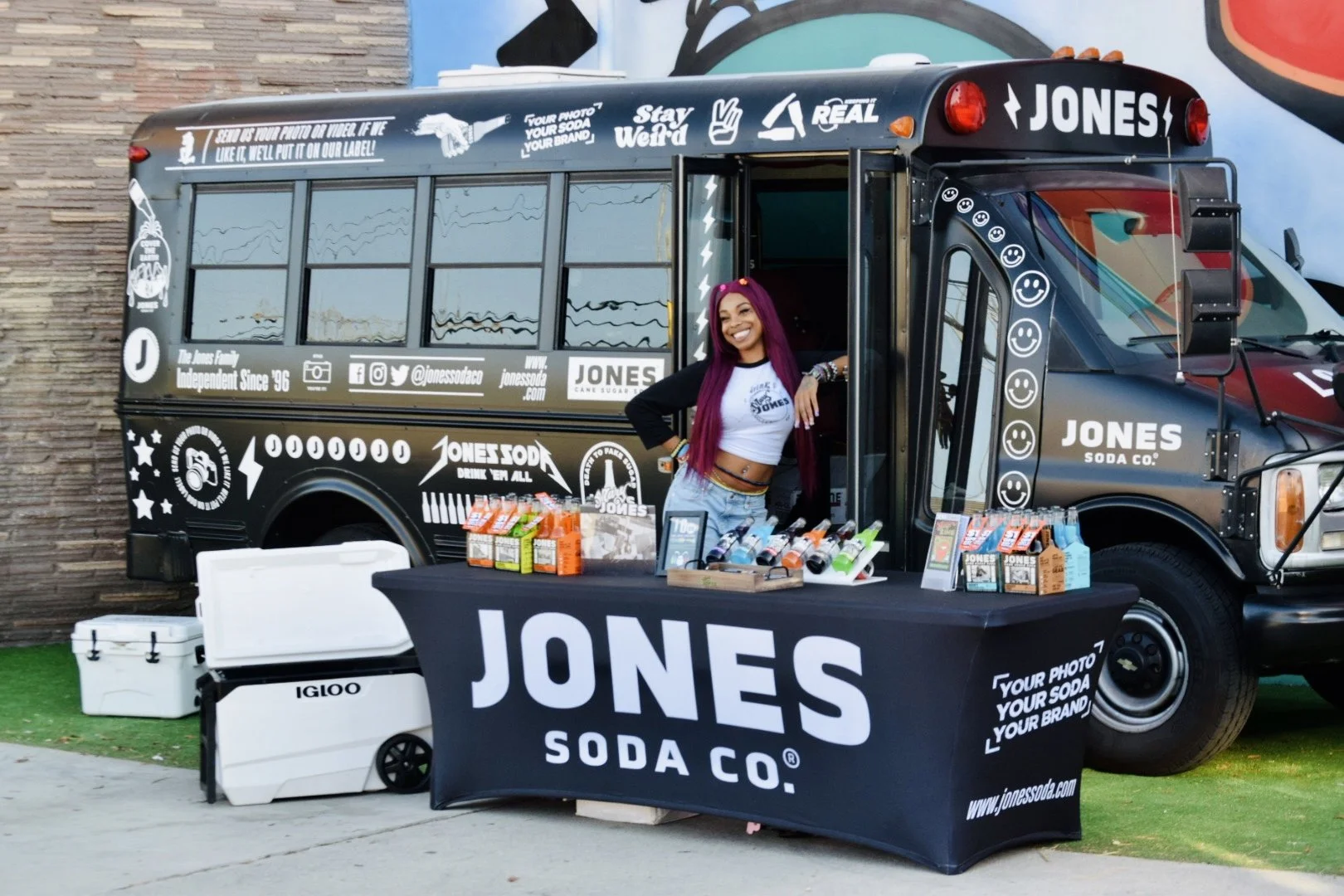 A woman with long pink hair smiling and posing in front of a Jones Soda Co. promotional bus, with a table displaying soda bottles and sampling products.