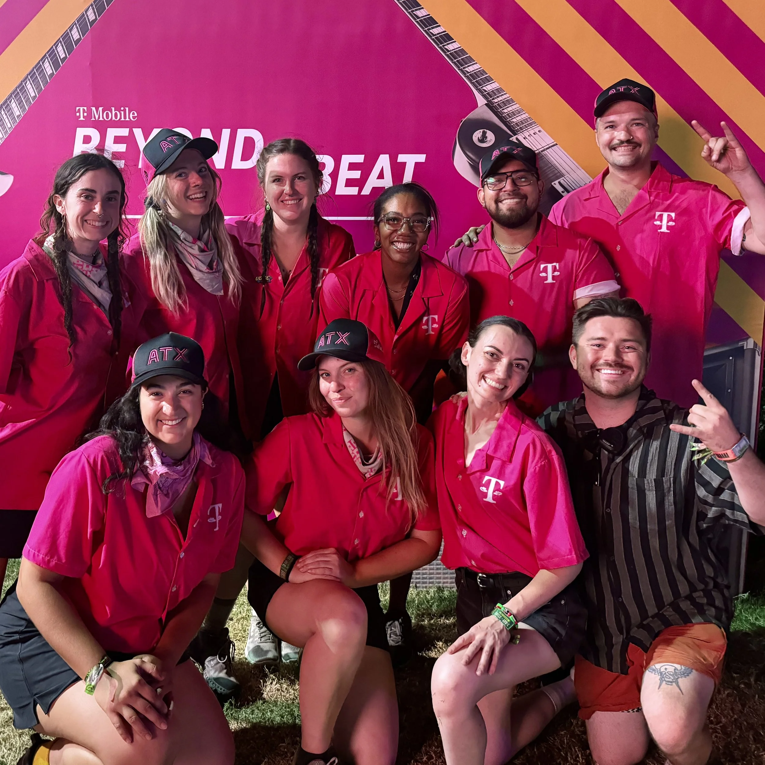 A group of six young adults wearing matching pink shirts and black hats with pink lettering, smiling and posing outdoors in front of a pink and yellow background with guitar graphics, at an event.