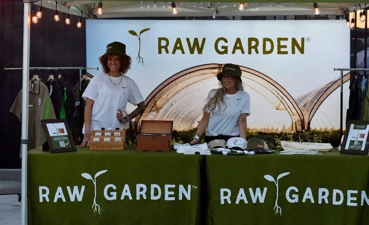 Two women standing behind a table at a RAW GARDEN booth, selling products and wearing white t-shirts and hats. The backdrop displays the RAW GARDEN logo with a sprouting plant, and the table has a green tablecloth with the same logo.