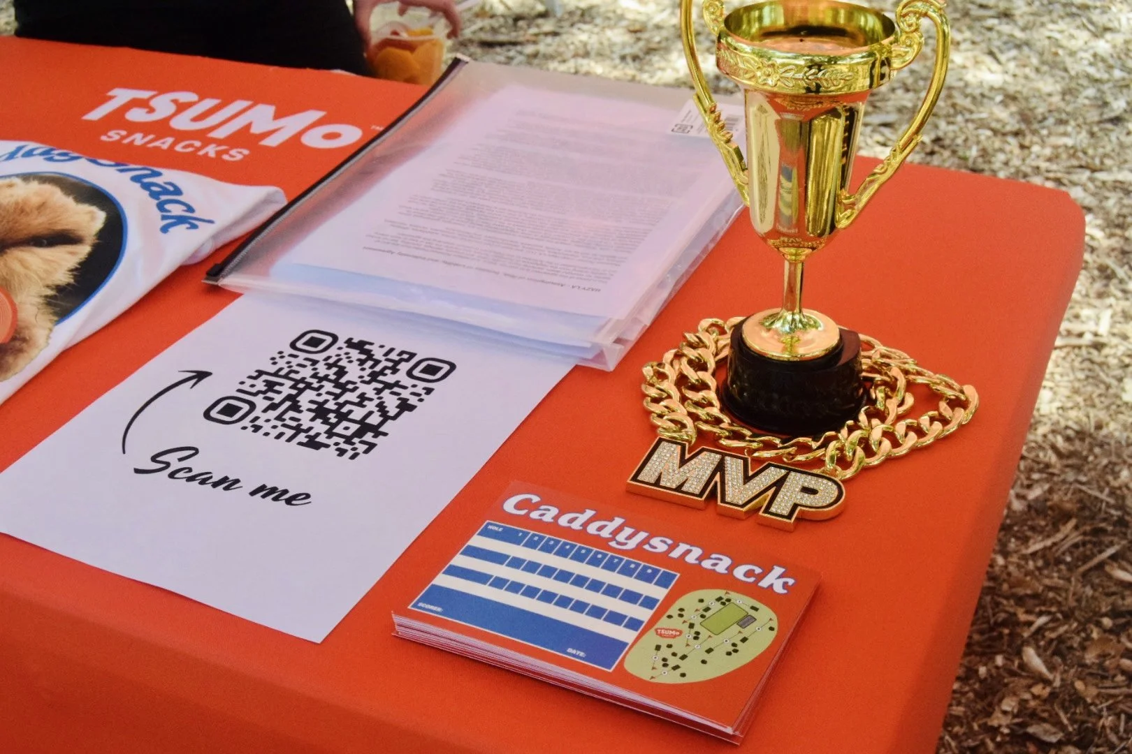 A gold trophy with handles and a large 'MVP' chain around its base, placed on an orange table. The table has a QR code sign that says 'Scan me,' a purple Caddy snack package, and some printed papers. The background shows a wilderness ground with leaves and twigs.