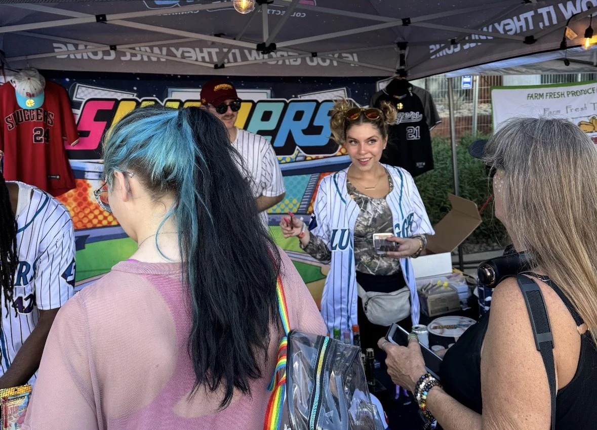 People at a stall under a tent at a fair or market, with a woman in a baseball jersey talking to customers, some merchandise like jerseys and hats displayed behind her.